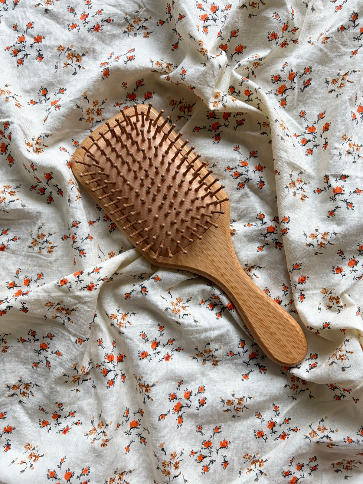 A wooden hairbrush with natural bamboo-like handles and bristles, placed on a white fabric background with a small orange and green floral print.