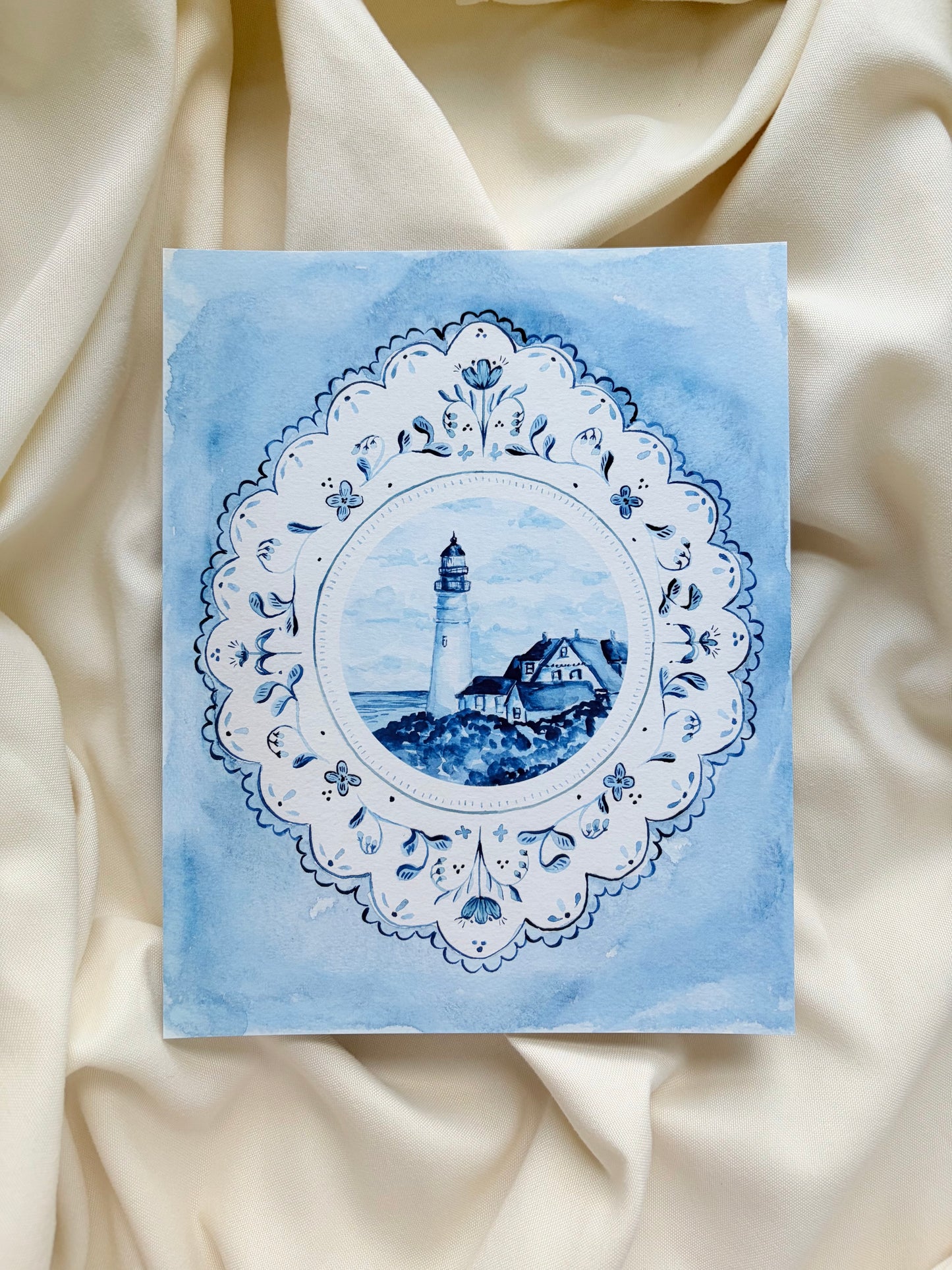 Decorative art print with lighthouse design on a blue background, placed on beige fabric.