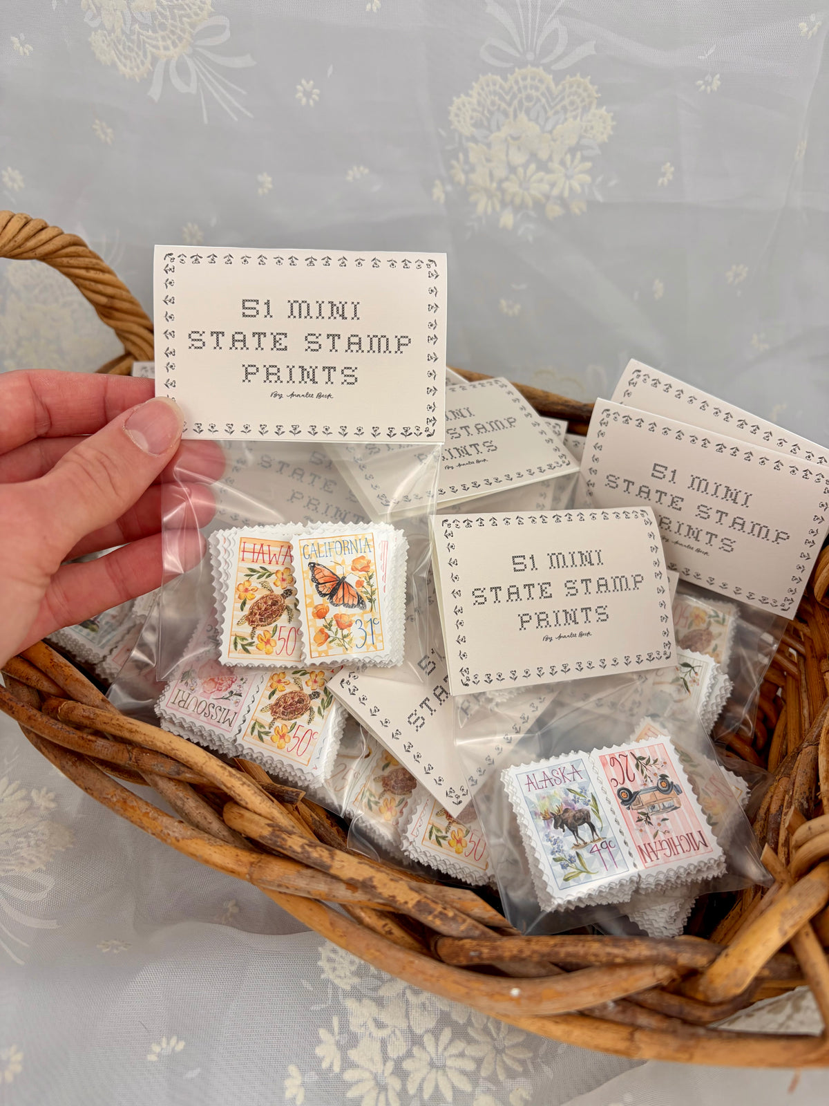 A hand holds a clear plastic bag containing a miniature stamp print, placed inside a woven wicker basket filled with multiple similar packaged mini stamp prints. Each package has a small card labeled '51 MINI STATE STAMP PRINTS.