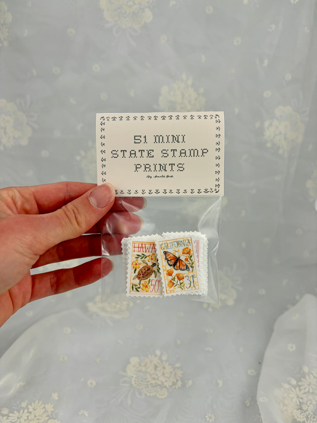 A hand holds a clear plastic bag containing a set of miniature state stamp prints with a paper label that reads '51 MINI STATE STAMP PRINTS by ditsy dot'. The background is a white lace curtain.