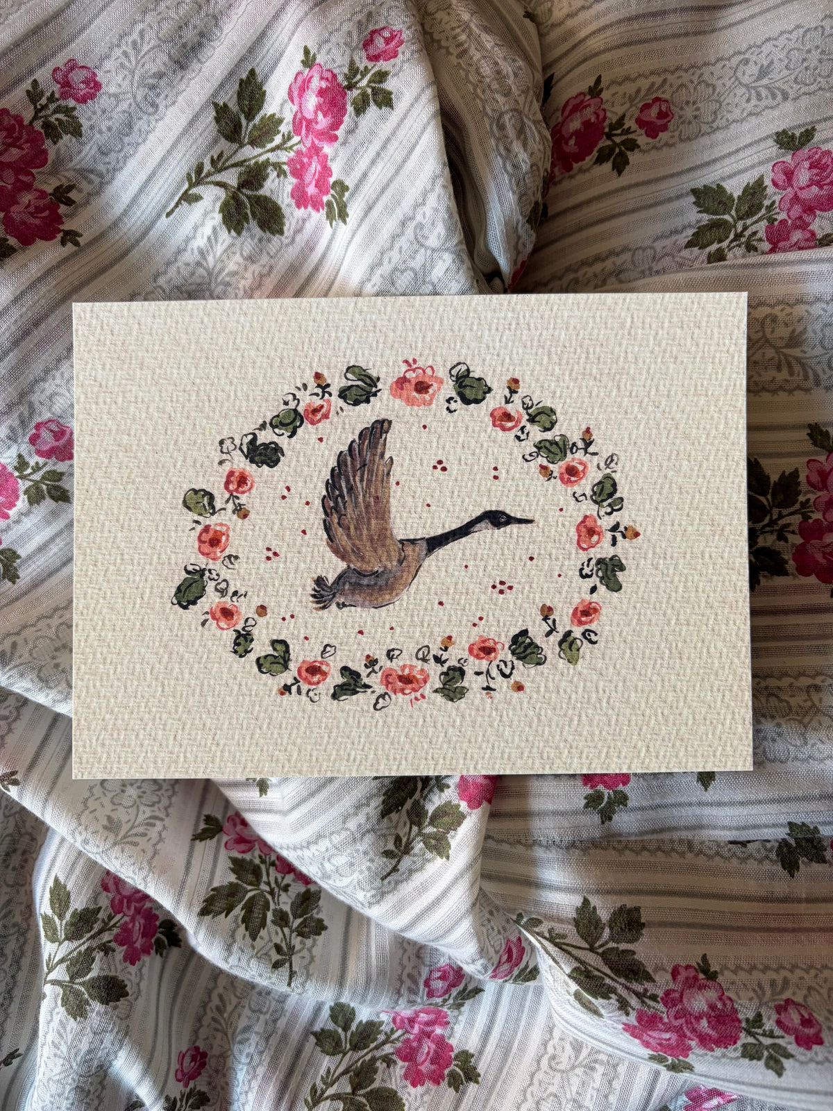 A square cream-colored textured print featuring an embroidered-style illustration of a flying goose surrounded by a floral wreath, laid flat on a patterned fabric background of pink roses and stripes.