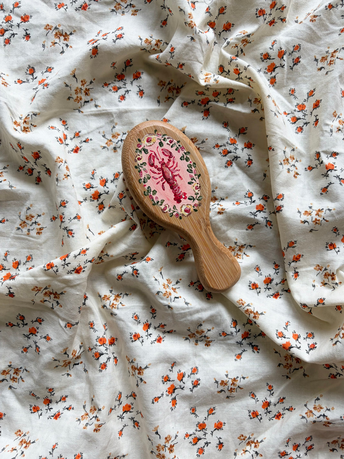 A wooden hairbrush with a pink lobster illustration and floral detailing, resting on a white fabric background with a small orange and green floral print.