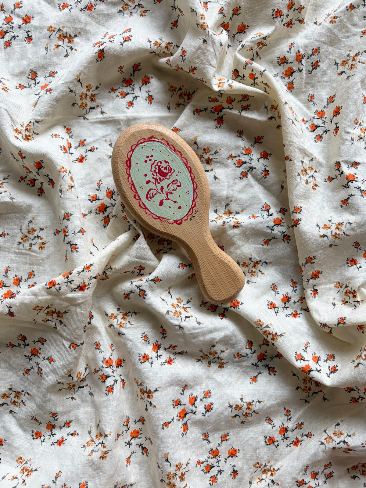 A small oval wooden hairbrush with a light blue/green center and red floral design, placed on rumpled white fabric with a small orange and black floral print.