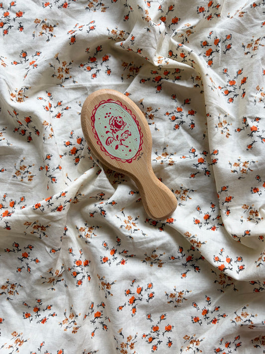 A small oval wooden hairbrush with a light blue/green center and red floral design, placed on rumpled white fabric with a small orange and black floral print.