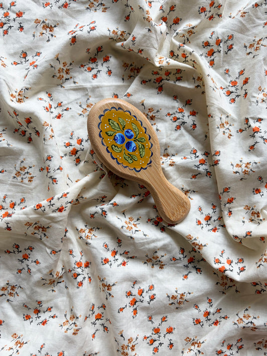 A small wooden hairbrush with a colorful hand-painted blueberry design, resting on a rumpled white fabric background with a small orange and black floral print.