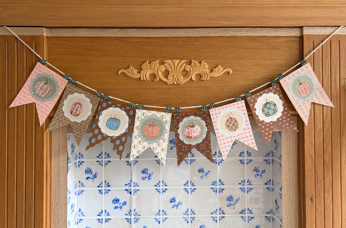 A decorative bunting banner with multiple patterned pennants in pink, brown, beige, and cream, each featuring a stylized pumpkin illustration and scalloped or patterned borders, hung across a wooden door above a blue-and-white tiled fireplace for styling purposes only.
