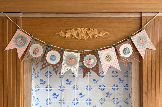 A decorative bunting banner with multiple patterned pennants in pink, brown, beige, and cream, each featuring a stylized pumpkin illustration and scalloped or patterned borders, hung across a wooden door above a blue-and-white tiled fireplace for styling purposes only.