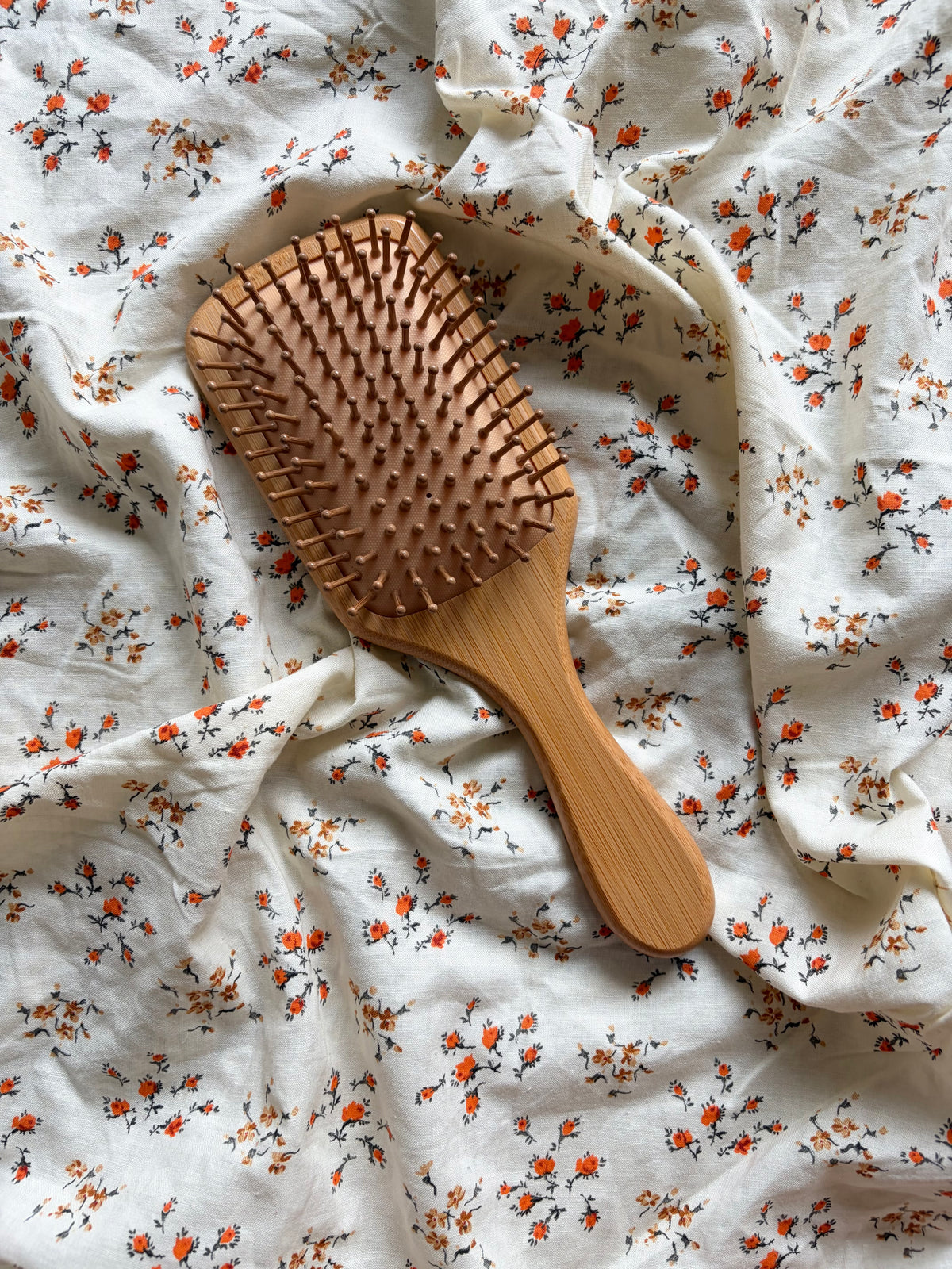 A wooden hairbrush with a warm brown finish, placed diagonally on a white fabric background with a small orange and green floral print.