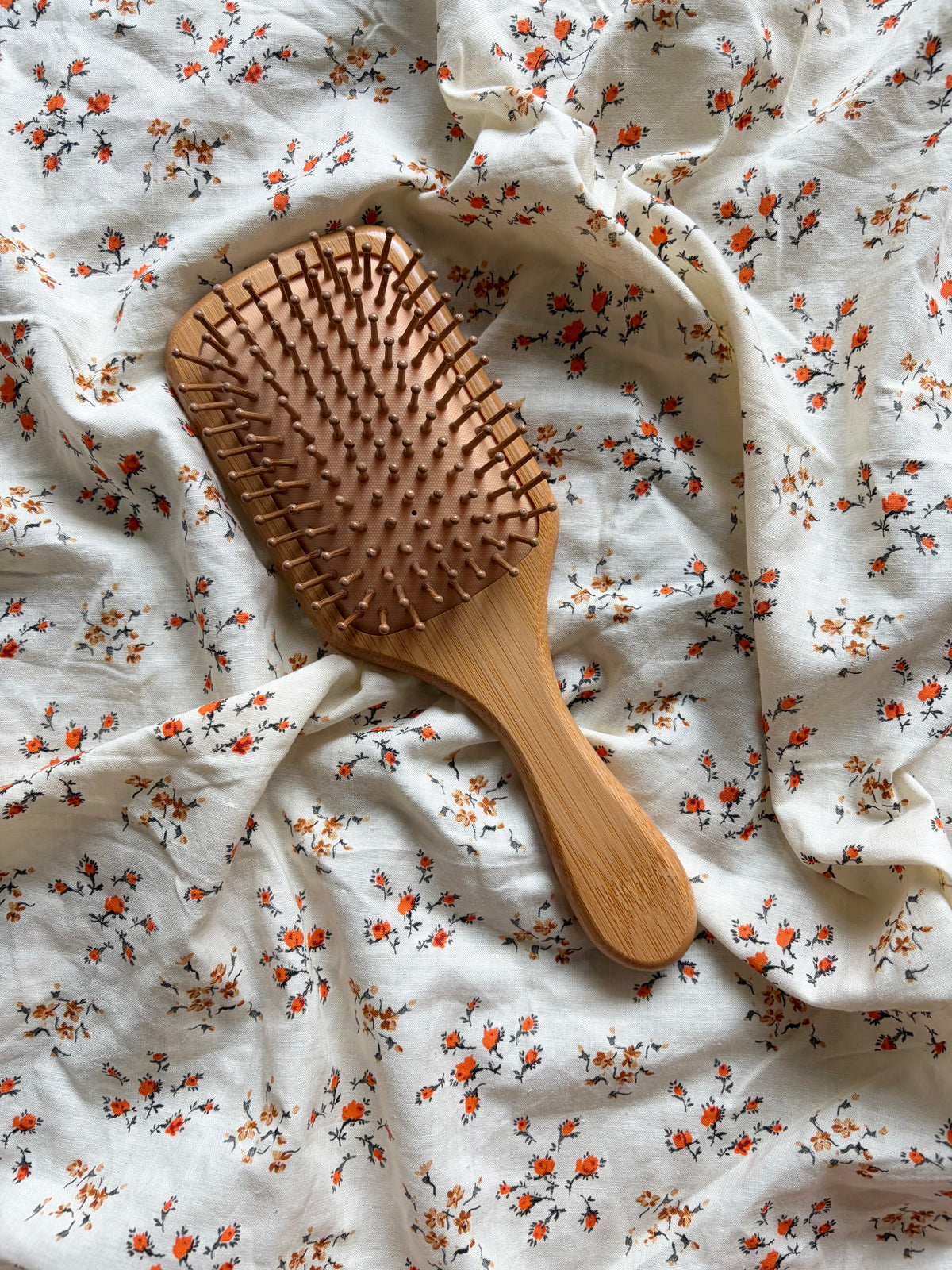 A wooden hairbrush with a natural bamboo finish, placed on a white fabric background with a small orange and green floral print.