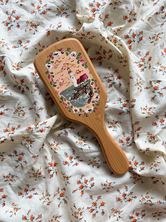A wooden hairbrush with a colorful lighthouse illustration, resting on a white fabric background with a small orange and green floral print.