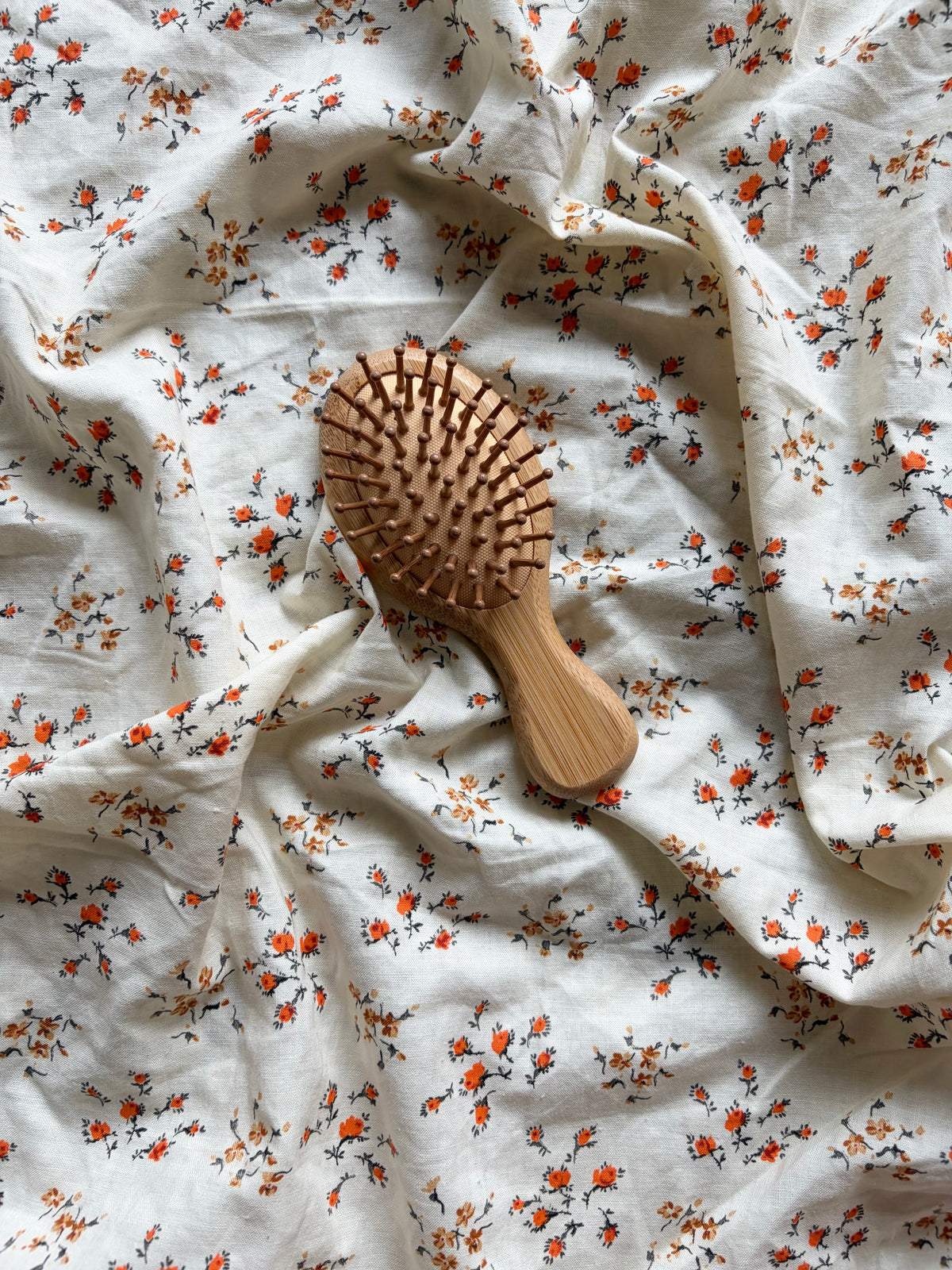 A wooden hairbrush with a light natural wood handle and bristles lies on rumpled white fabric printed with small orange and brown floral motifs.