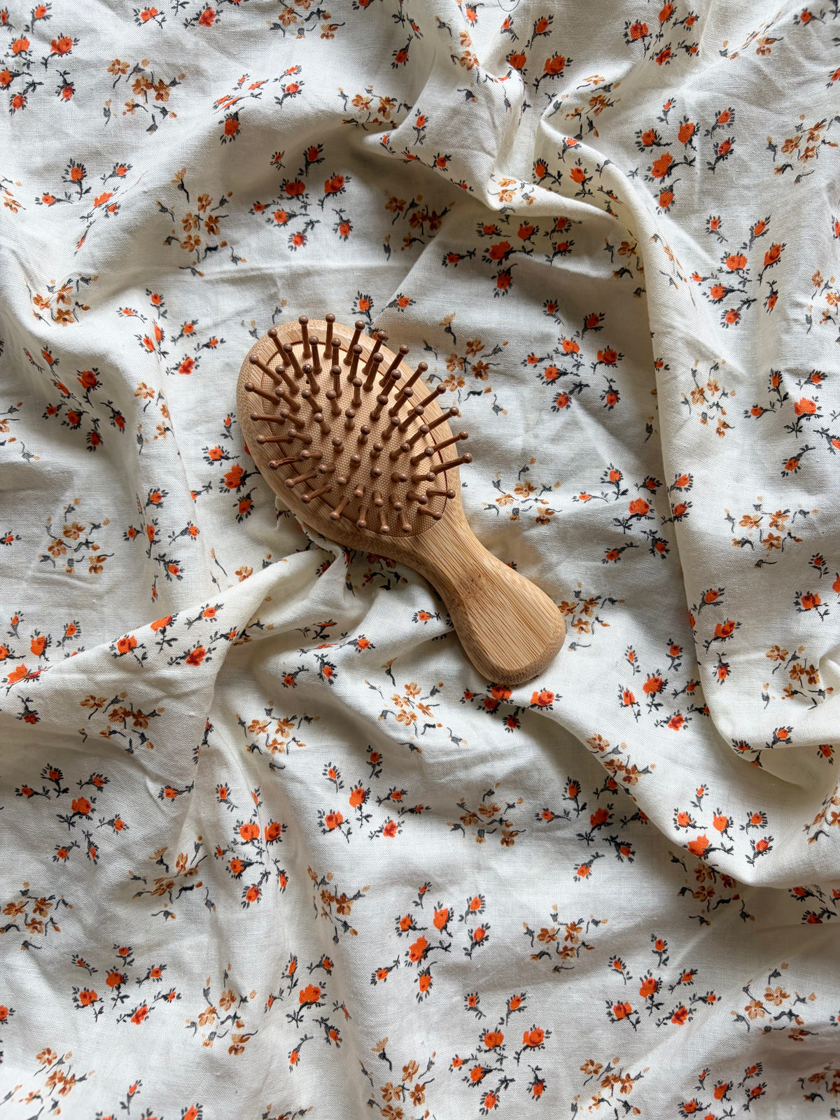 A wooden hairbrush with a light natural wood handle and bristles, placed on a draped white fabric background with a small orange and gray floral print.