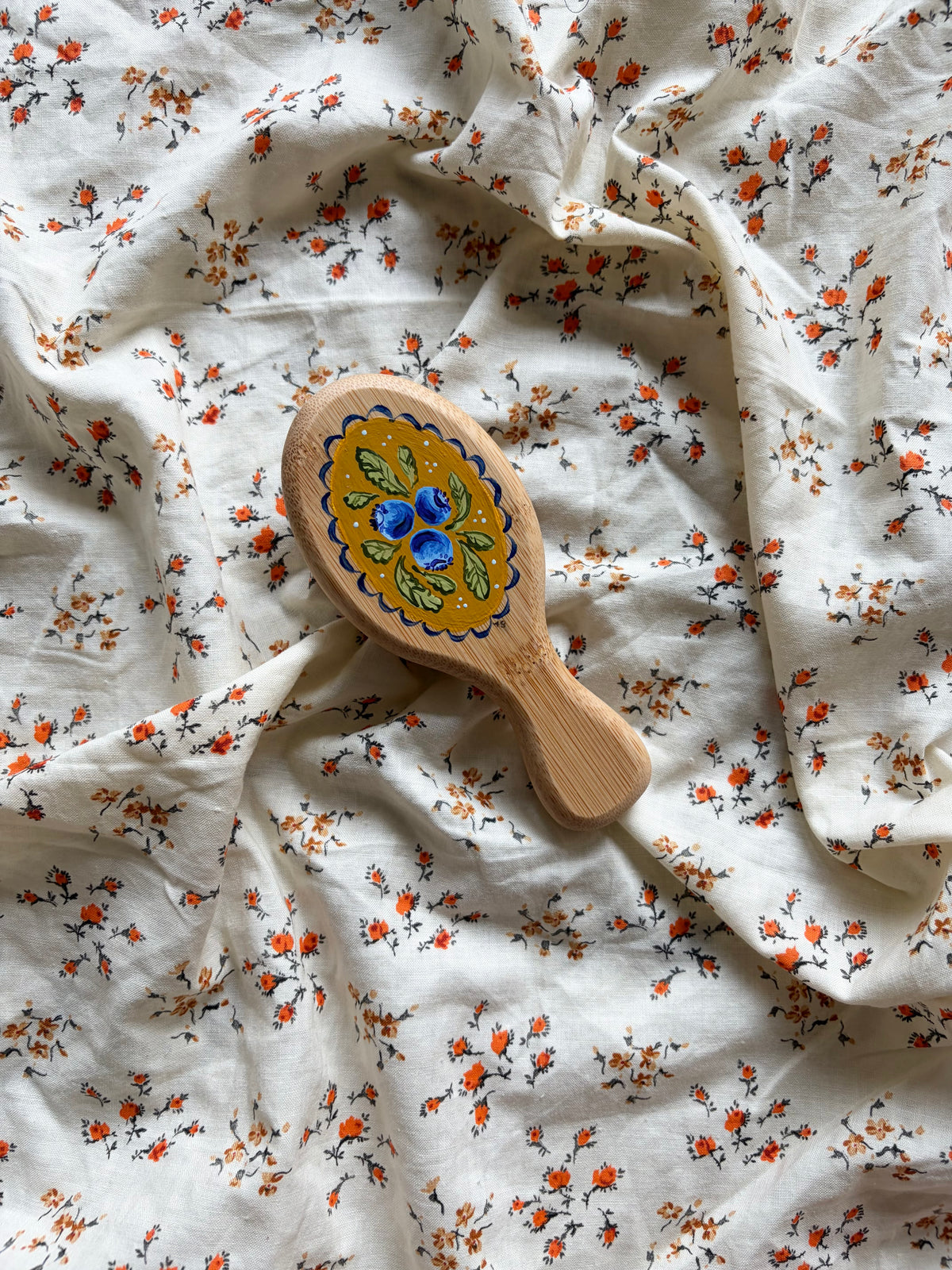 A small wooden hairbrush with a colorful hand-painted blueberry design, resting on a rumpled white fabric background with a small orange and black floral print.