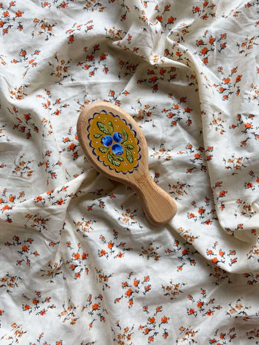 A small wooden hairbrush with a colorful hand-painted blueberry design, resting on a rumpled white fabric background with a small orange and black floral print.
