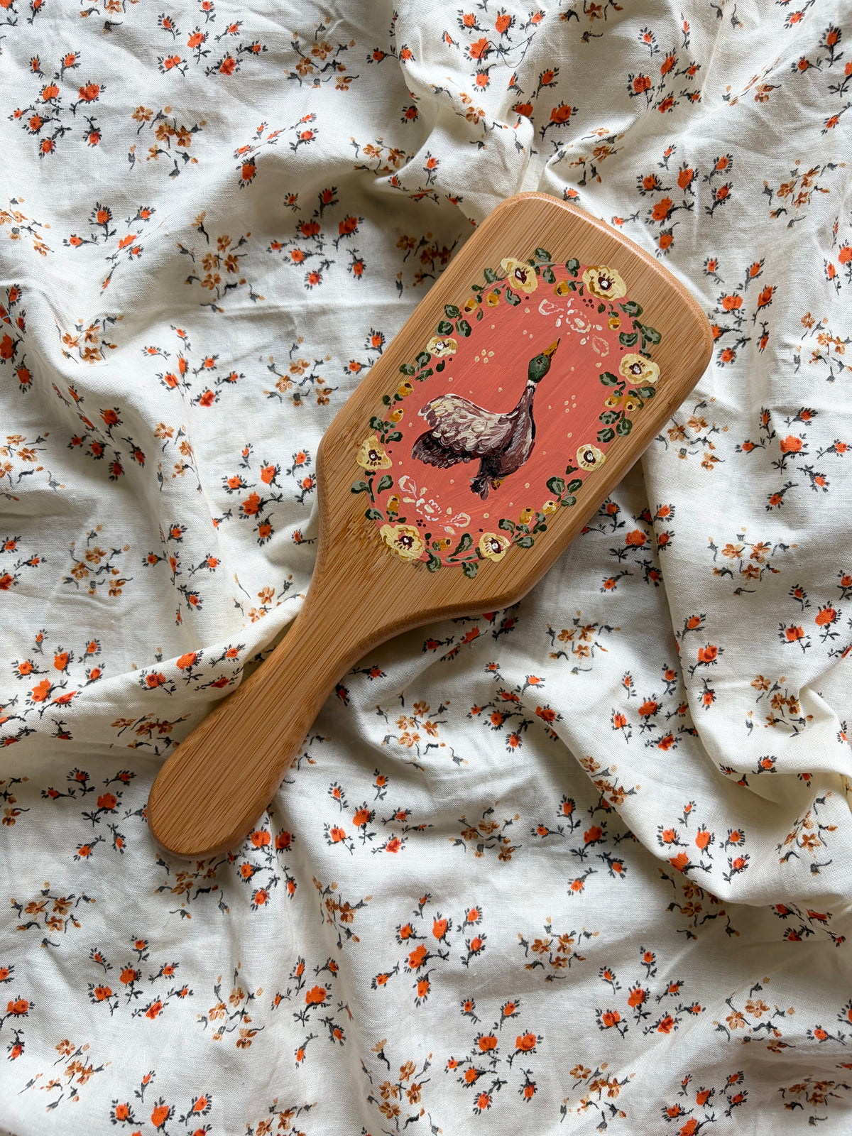 A wooden paddle hairbrush with a hand-painted mallard duck design on a coral background, surrounded by a floral border, laid diagonally on a white fabric with orange and green floral print.