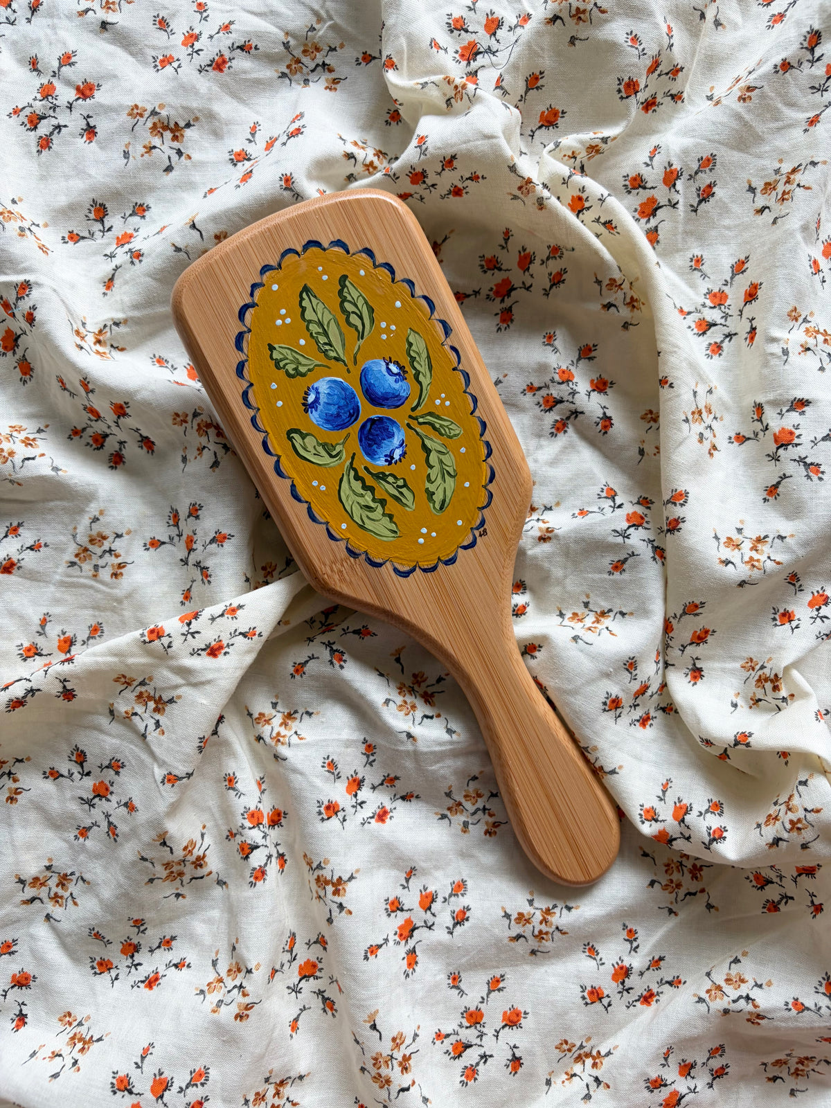 A wooden hairbrush with a hand-painted yellow, green, and blueberries design, resting on a white fabric background with a small orange floral print.