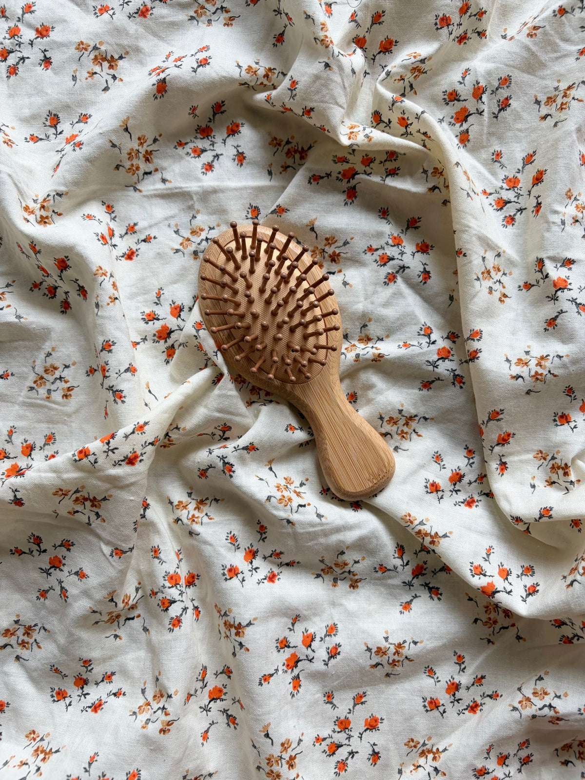 A wooden hairbrush with light brown wood and orange-brown bristles, placed on a draped white fabric background with a small orange and green floral print.