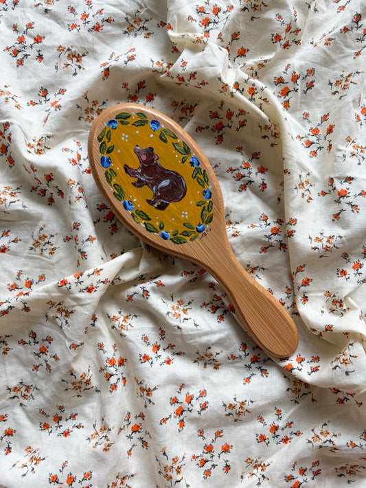 A wooden hairbrush with a painted brown bear and blueberry border, resting on a white fabric background with a small orange and green floral print.