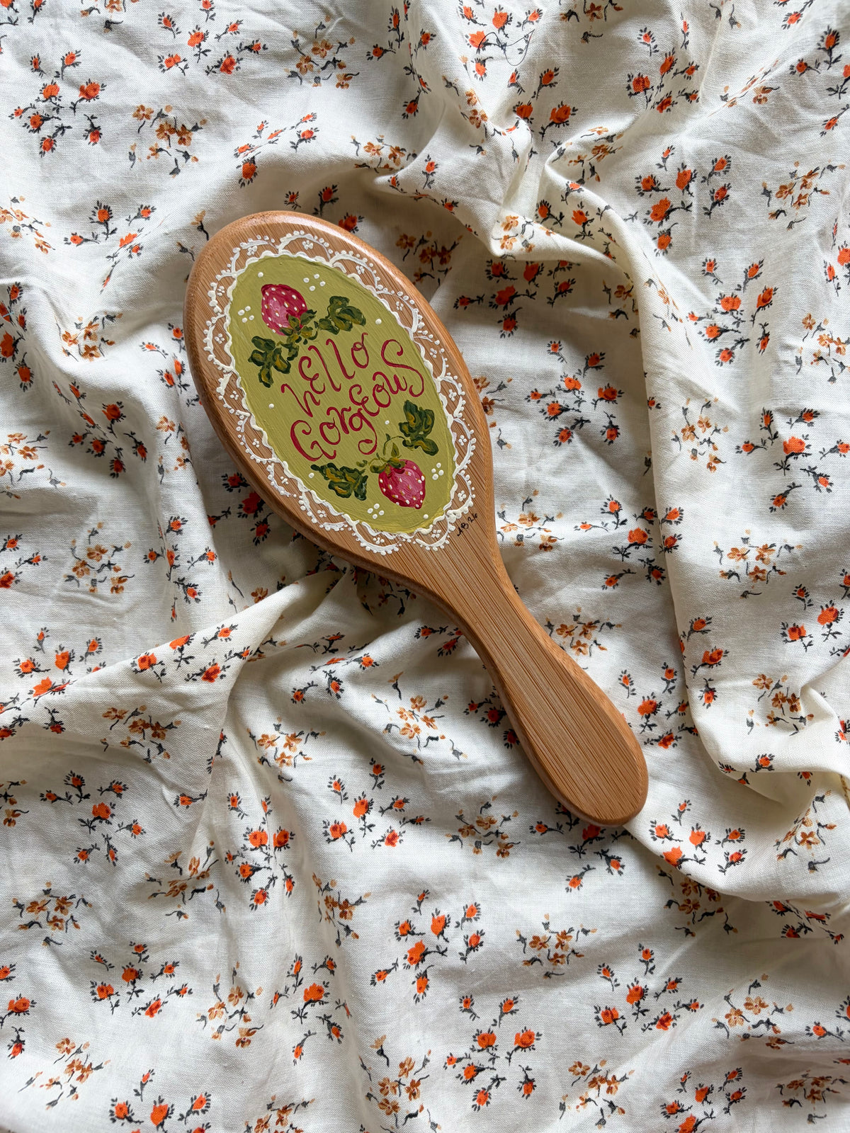 A wooden hairbrush with a decorative oval head lies on rumpled white fabric printed with small orange and green floral motifs. The brush is hand-painted with strawberries, green leaves, and the words 'Hello Gorgeous' in script.