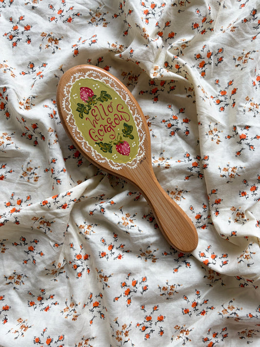 A wooden hairbrush with a decorative oval head lies on rumpled white fabric printed with small orange and green floral motifs. The brush is hand-painted with strawberries, green leaves, and the words 'Hello Gorgeous' in script.