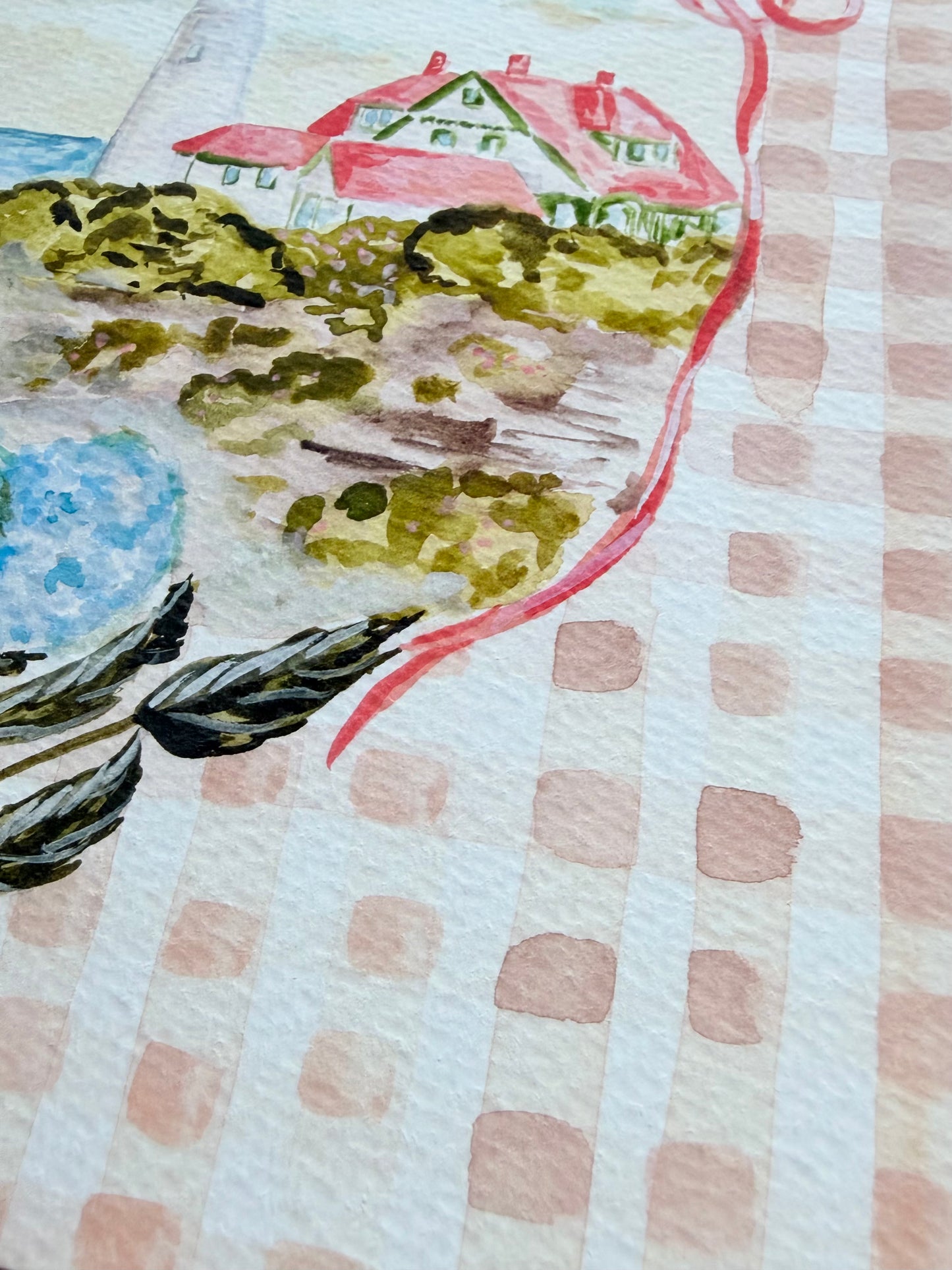 Close up photo of artwork with a landscape design featuring a lighthouse, bushes, and a blue sky on a gingham background.