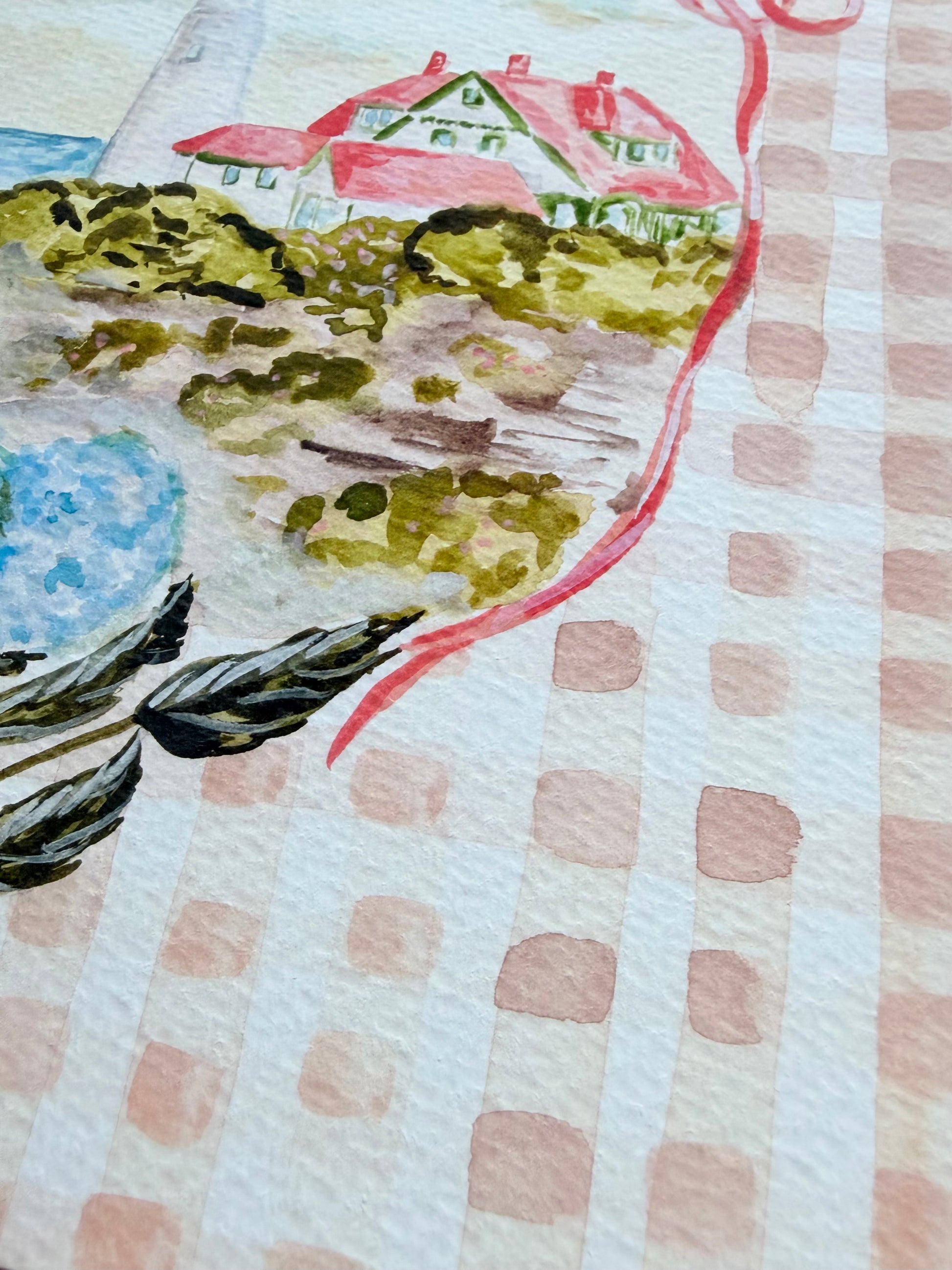 Close up photo of artwork with a landscape design featuring a lighthouse, bushes, and a blue sky on a gingham background.