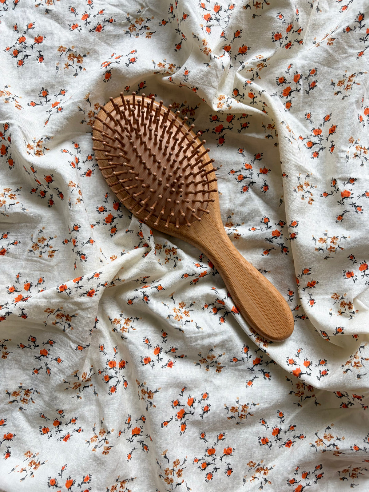 A wooden hairbrush with a smooth bamboo handle, placed diagonally on a rumpled white fabric background with a small orange and brown floral print.