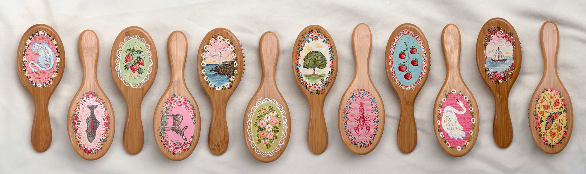 A row of 12 wooden hairbrushes with colorful, hand-painted floral and landscape designs on the brush heads, arranged on a white textured fabric background.