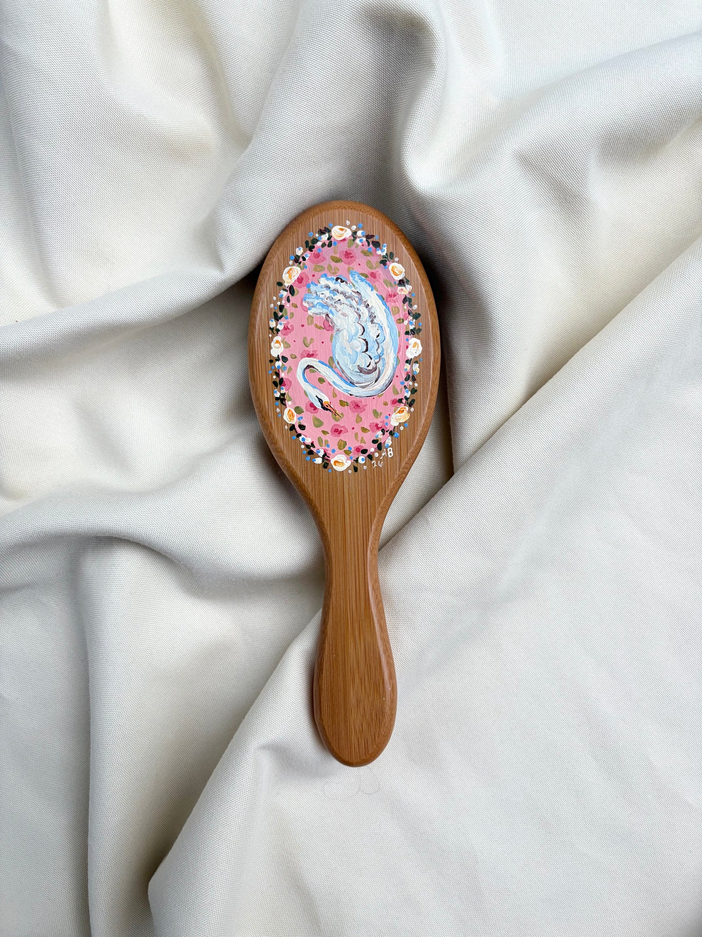 A wooden hairbrush with a pink floral and swan design on the oval head, resting on draped white fabric.