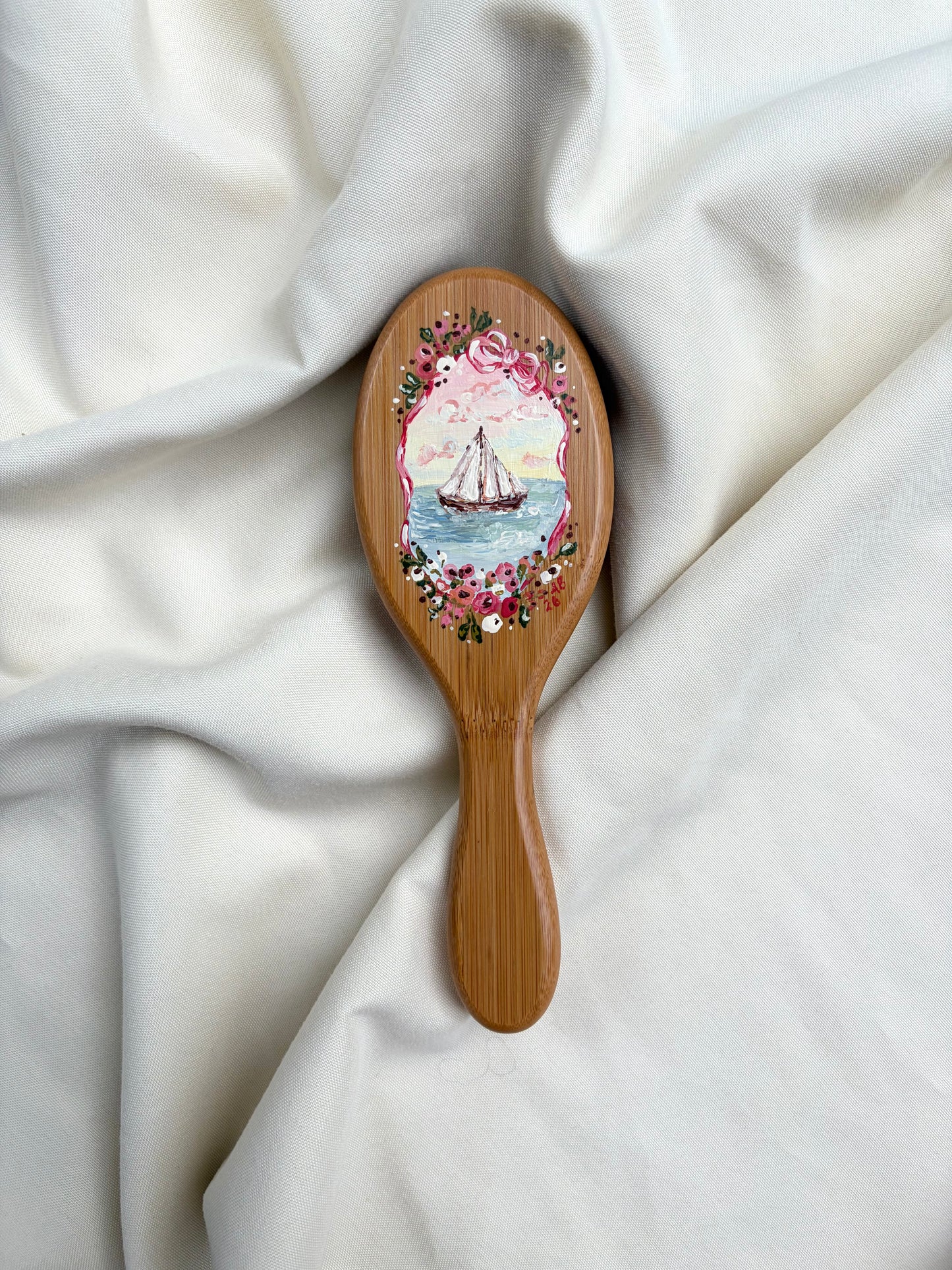 A wooden hairbrush with an oval head featuring a painted sailboat scene surrounded by pink flowers, resting on draped white fabric.