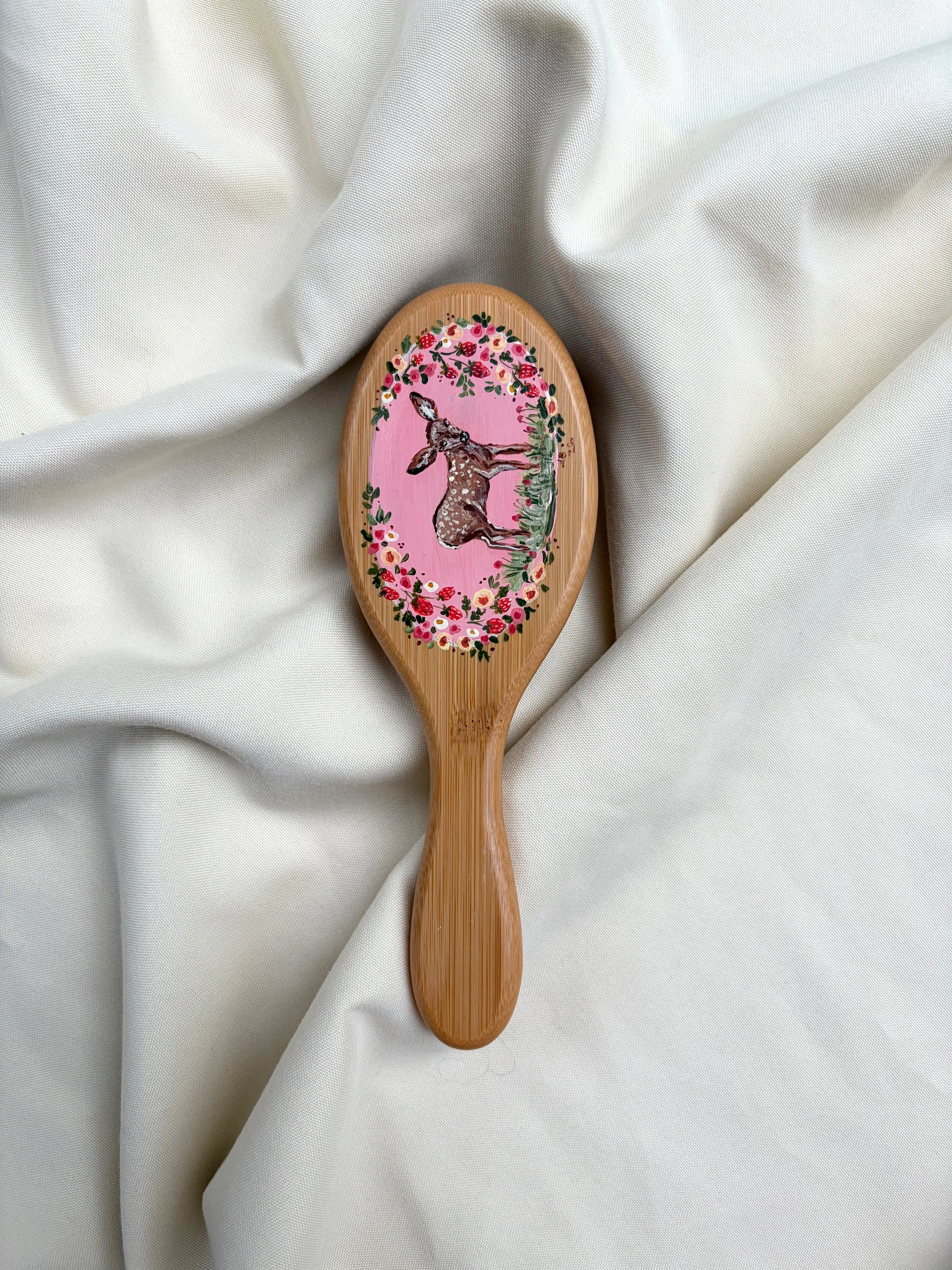 A wooden hairbrush with an oval head face featuring a deer illustration surrounded by a floral and strawberry wreath, resting on draped white fabric.