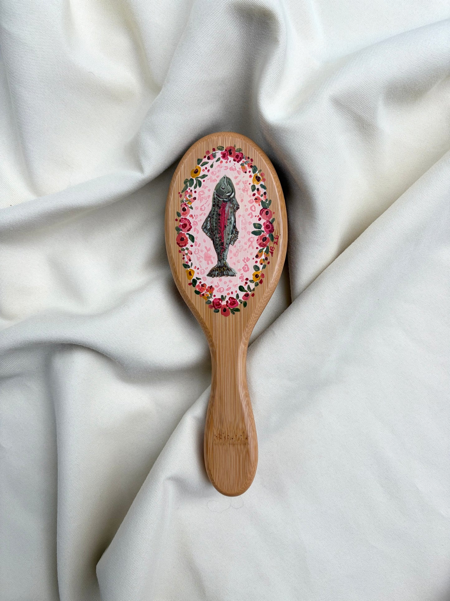 A wooden hairbrush with a pink floral oval design and a painted fish design with floral outline, resting on draped white fabric.