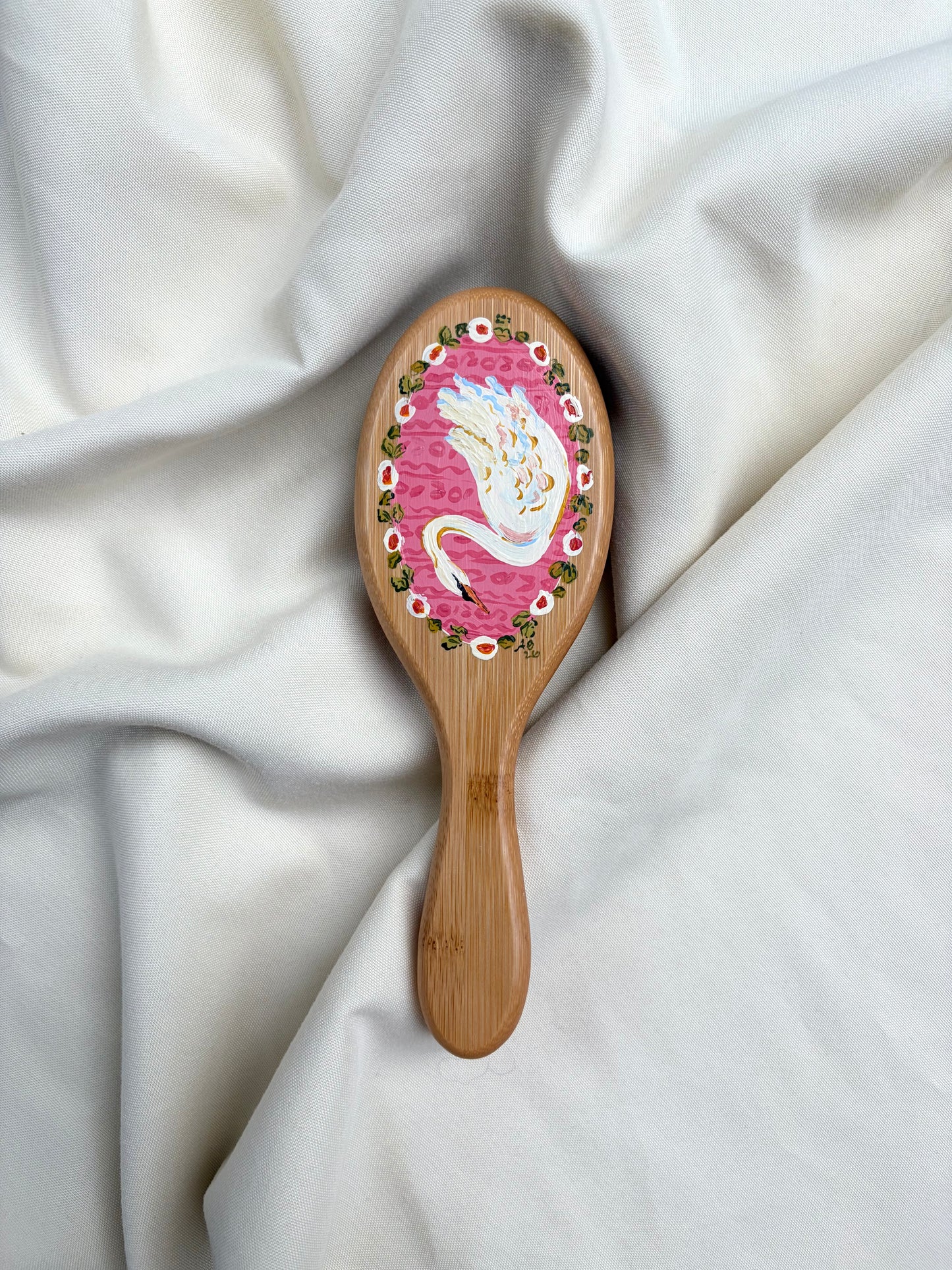A wooden hairbrush with a pink and white swan illustration on the brush head with florals, resting on draped white fabric.