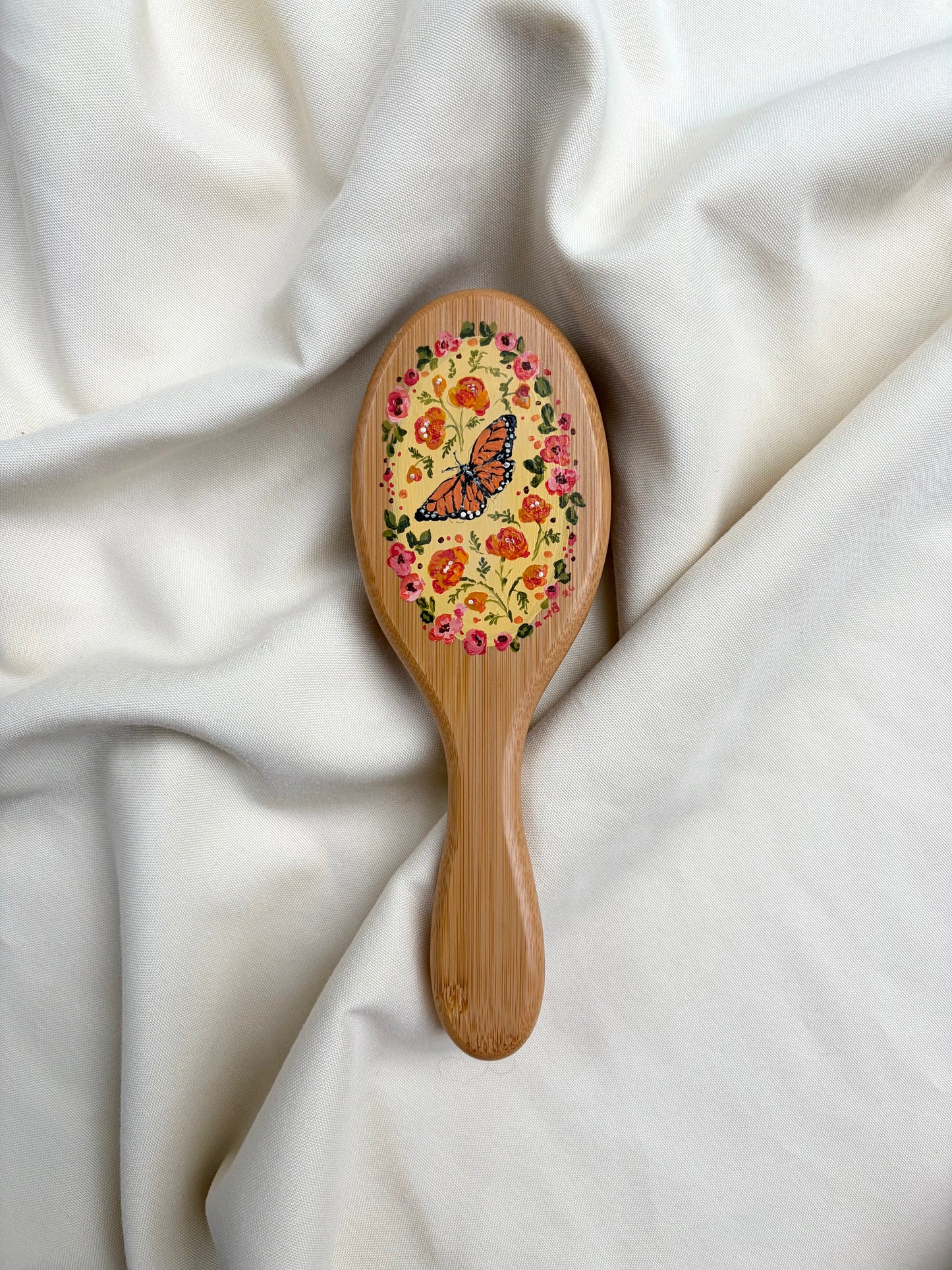 A small wooden hairbrush with a colorful floral and butterfly design on the oval head, resting on draped white fabric.
