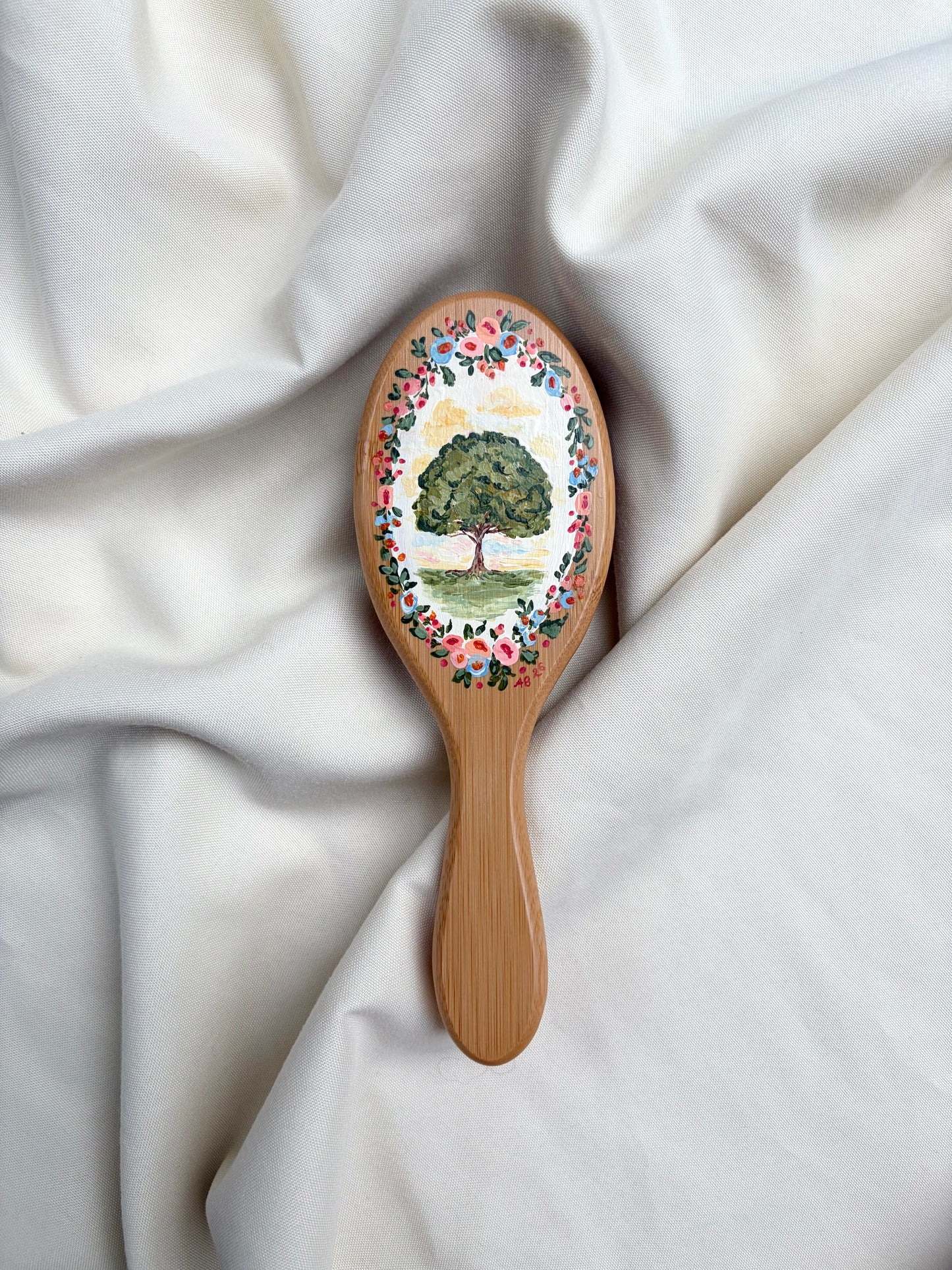 A wooden hairbrush with a hand-painted tree and floral design on the oval head, resting on draped white fabric.