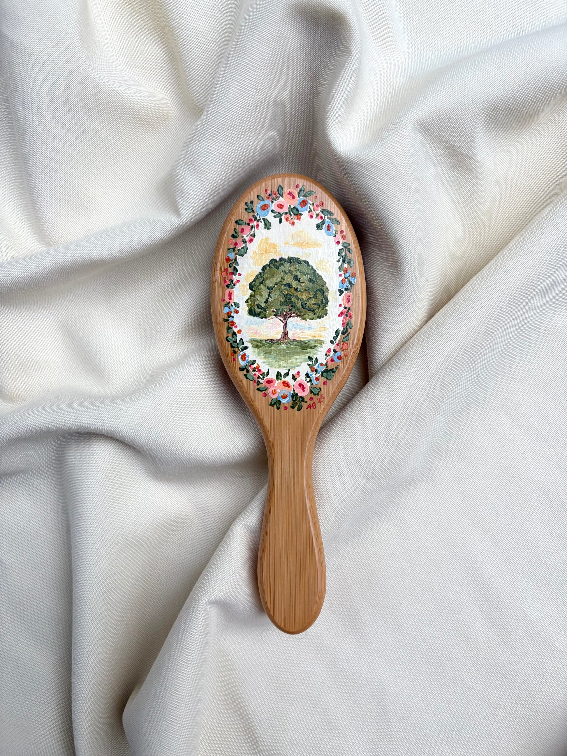 A wooden hairbrush with a hand-painted tree and floral design on the oval head, resting on draped white fabric.