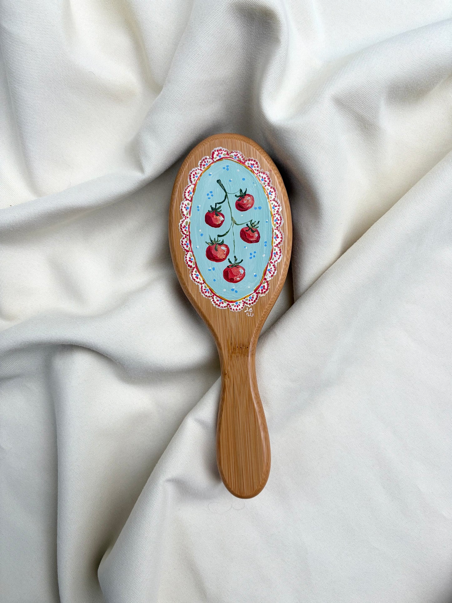 A wooden hairbrush with an oval handpainted design featuring a light blue background, red tomatoes, and a red and yellow floral border, resting on draped white fabric.