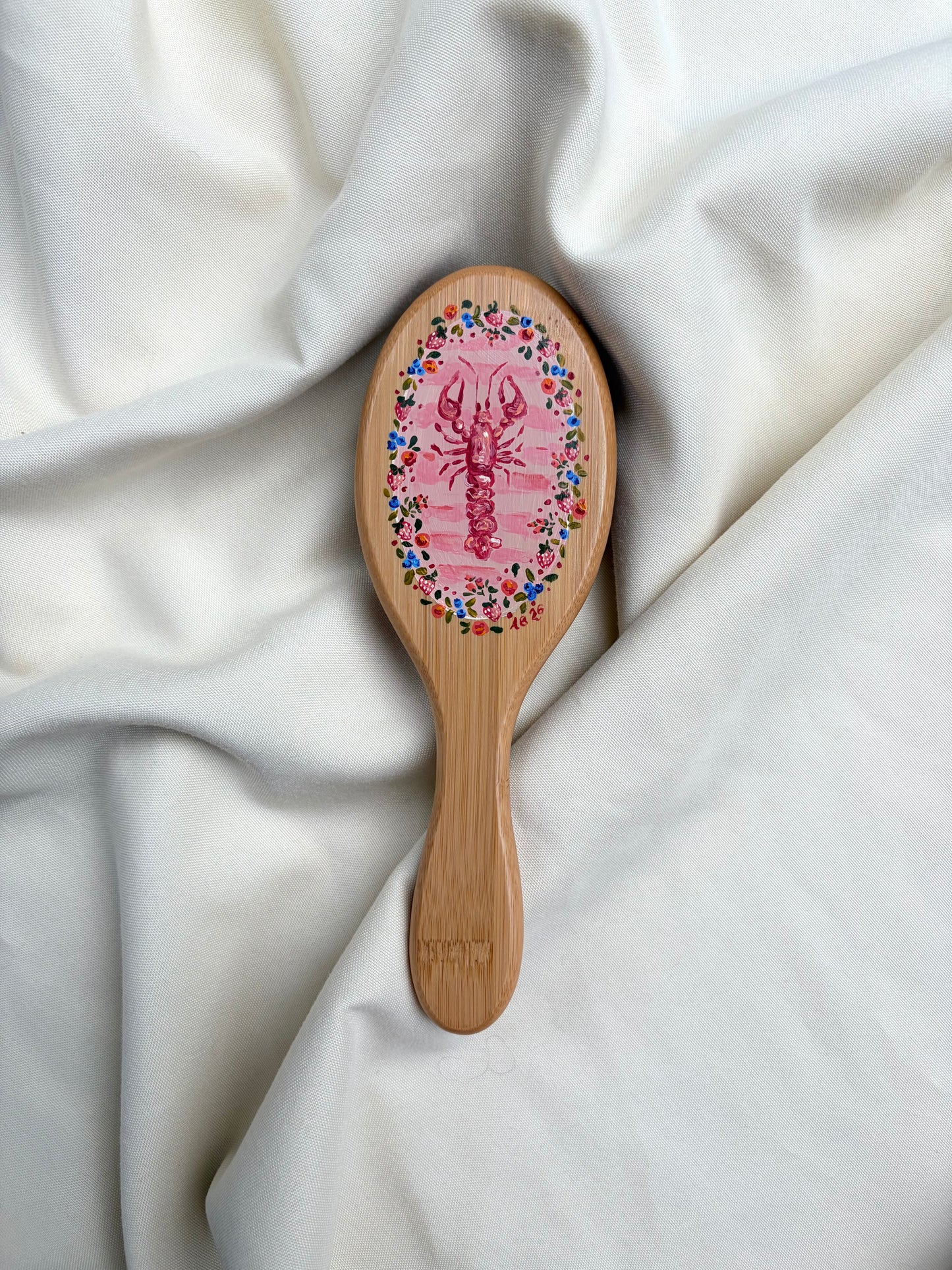 A bamboo hairbrush with a pink lobster illustration and a colorful floral border on the brush head, resting on draped white fabric.