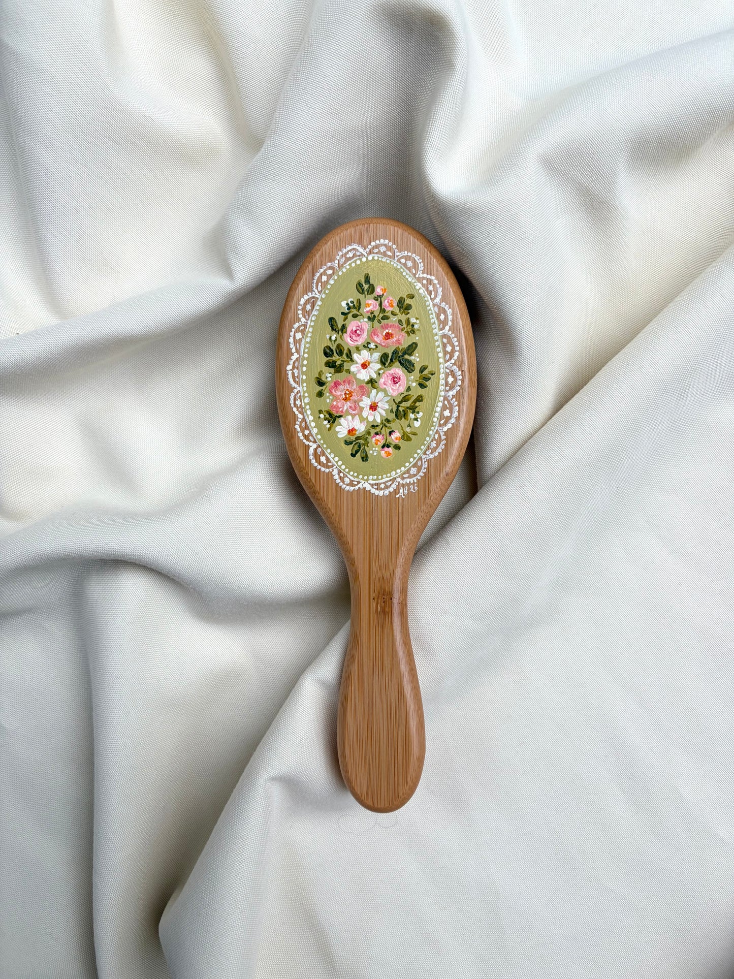 A wooden hairbrush with an oval face decorated with a floral design and white lace border, resting on draped white fabric.