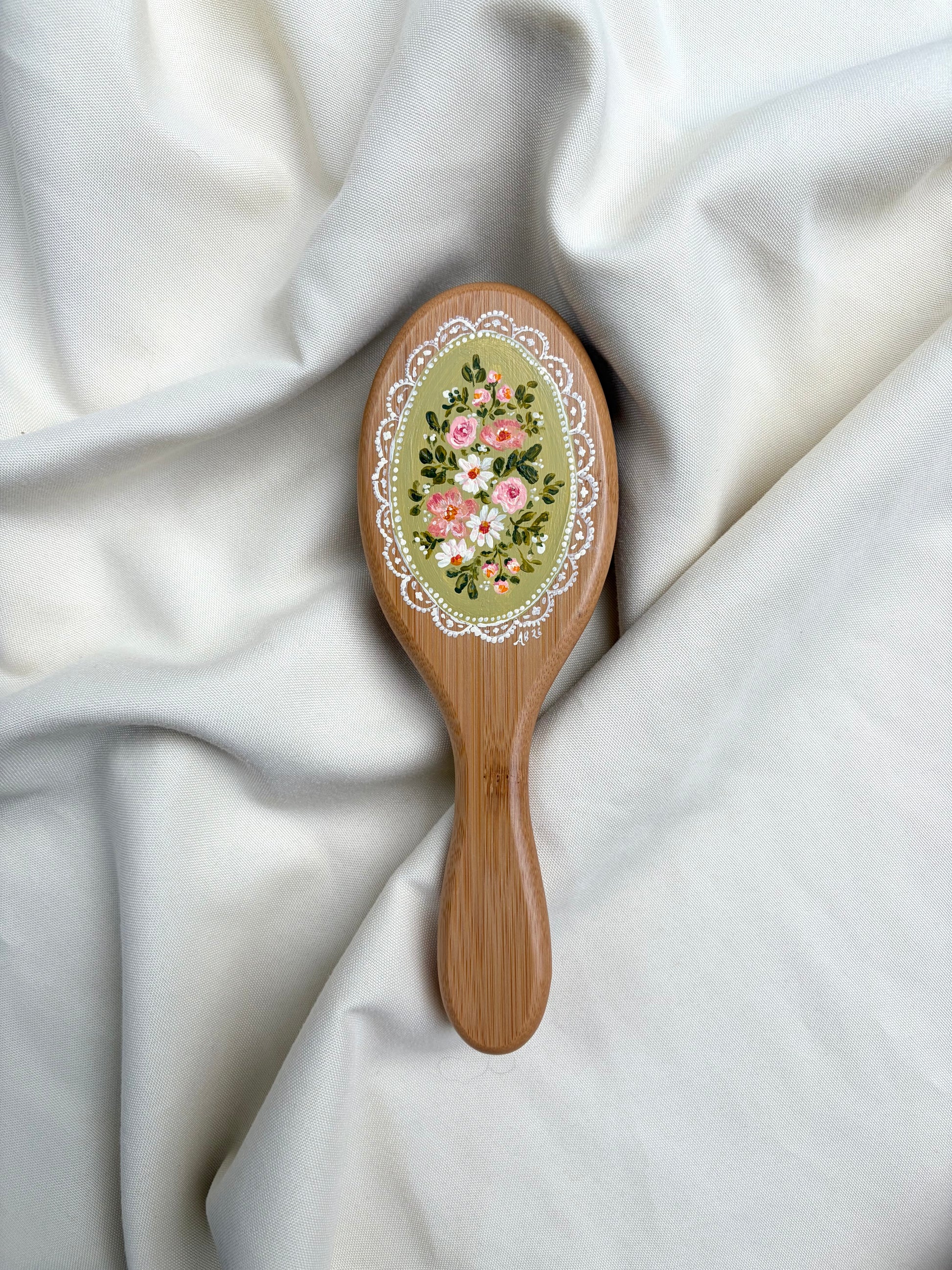 A wooden hairbrush with an oval face decorated with a floral design and white lace border, resting on draped white fabric.