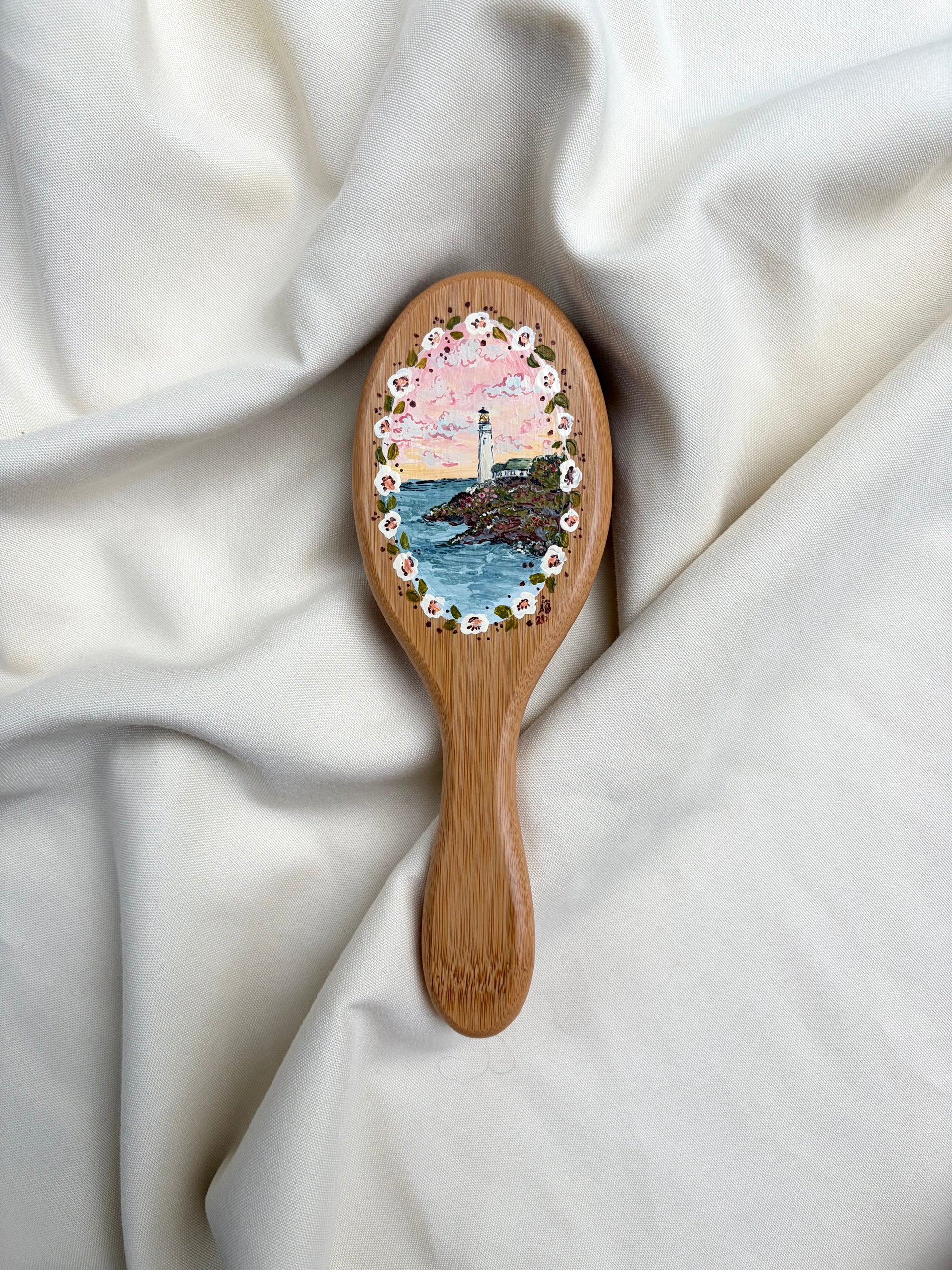 A wooden hairbrush with an oval painted part featuring a colorful lighthouse and ocean scene, surrounded by a floral border, resting on draped white fabric.