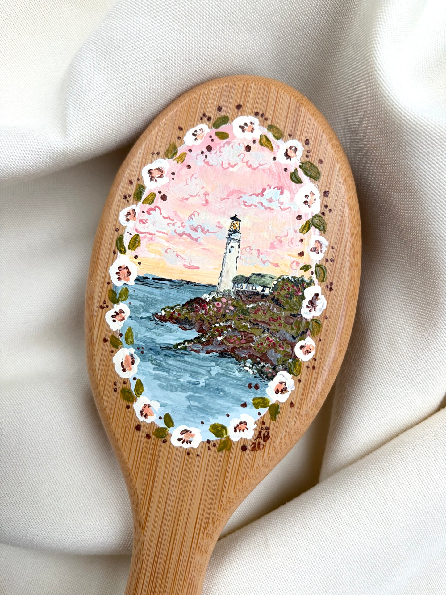 A hairbrush with a hand-painted lighthouse scene in the center, surrounded by a floral border, resting on draped cream fabric.