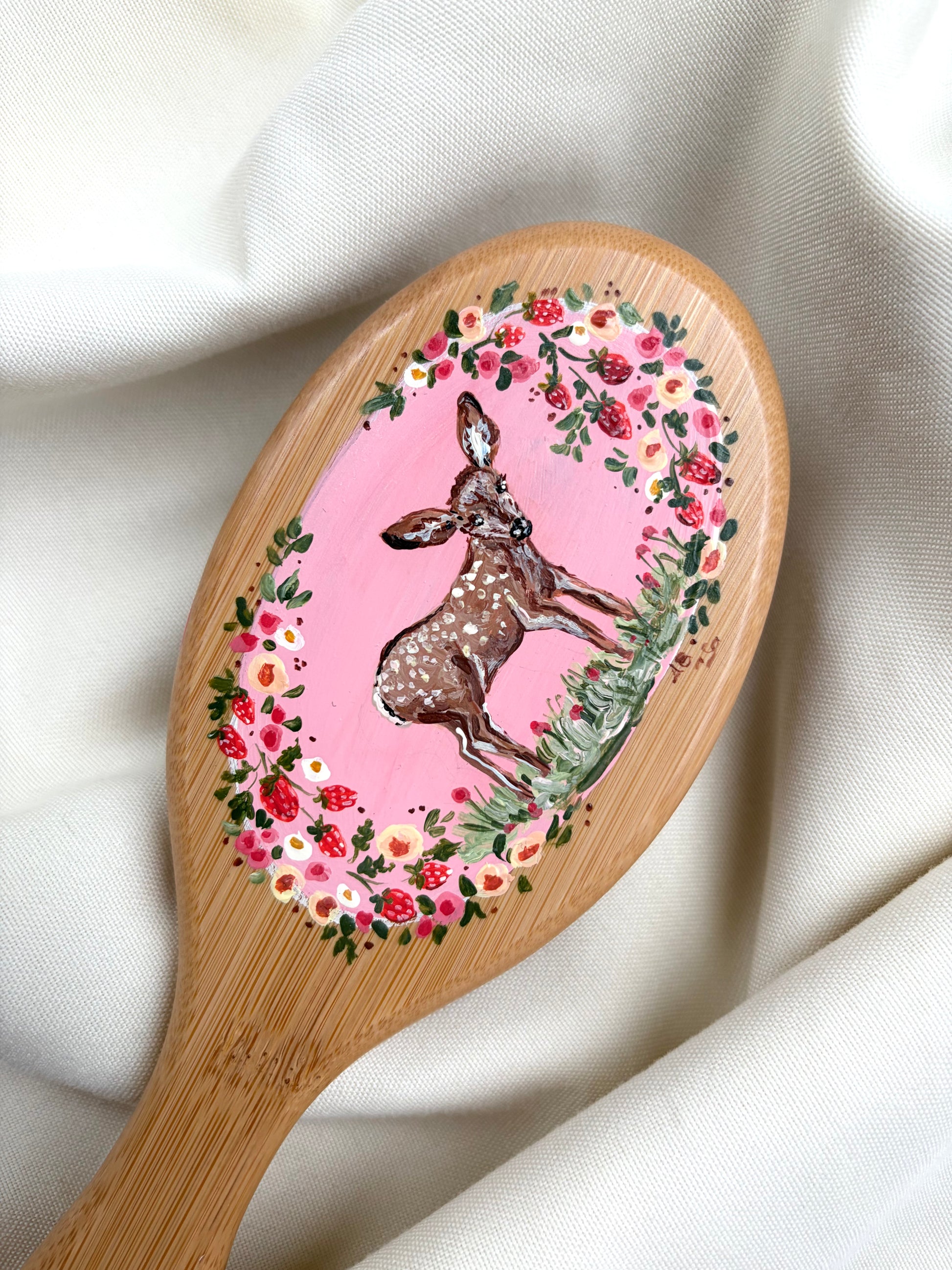 A wooden hairbrush with a natural bamboo handle and a painted oval head, featuring a brown deer surrounded by a pink floral and strawberry wreath, displayed on draped white fabric.