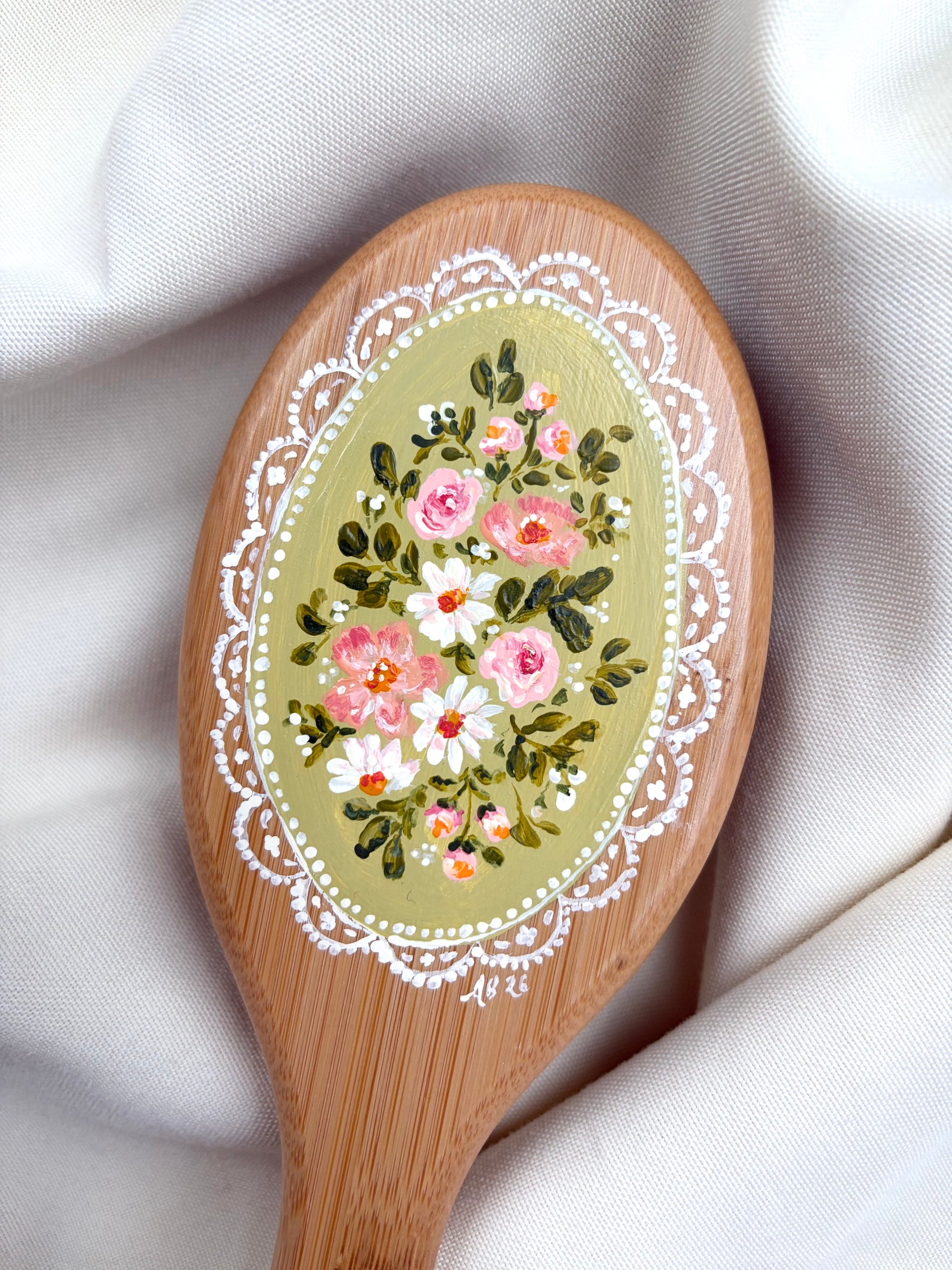 A close-up of a wooden hairbrush with an oval hand-painted floral design in pink, white, and green on a light wood, resting on draped cream fabric.