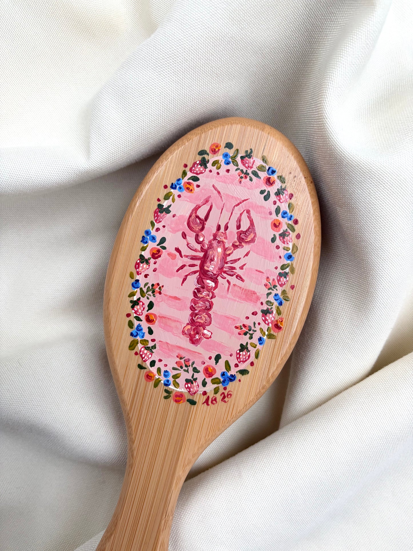 A close-up of a wooden hairbrush with a hand-painted red lobster in the center, surrounded by a colorful floral border, resting on draped white fabric.