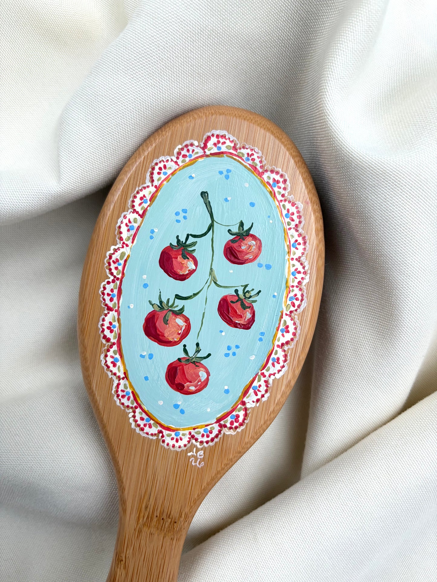 A close-up of a brush with a hand-painted design featuring red tomatoes on a light blue background, bordered by a white and red floral pattern, resting on draped white fabric.