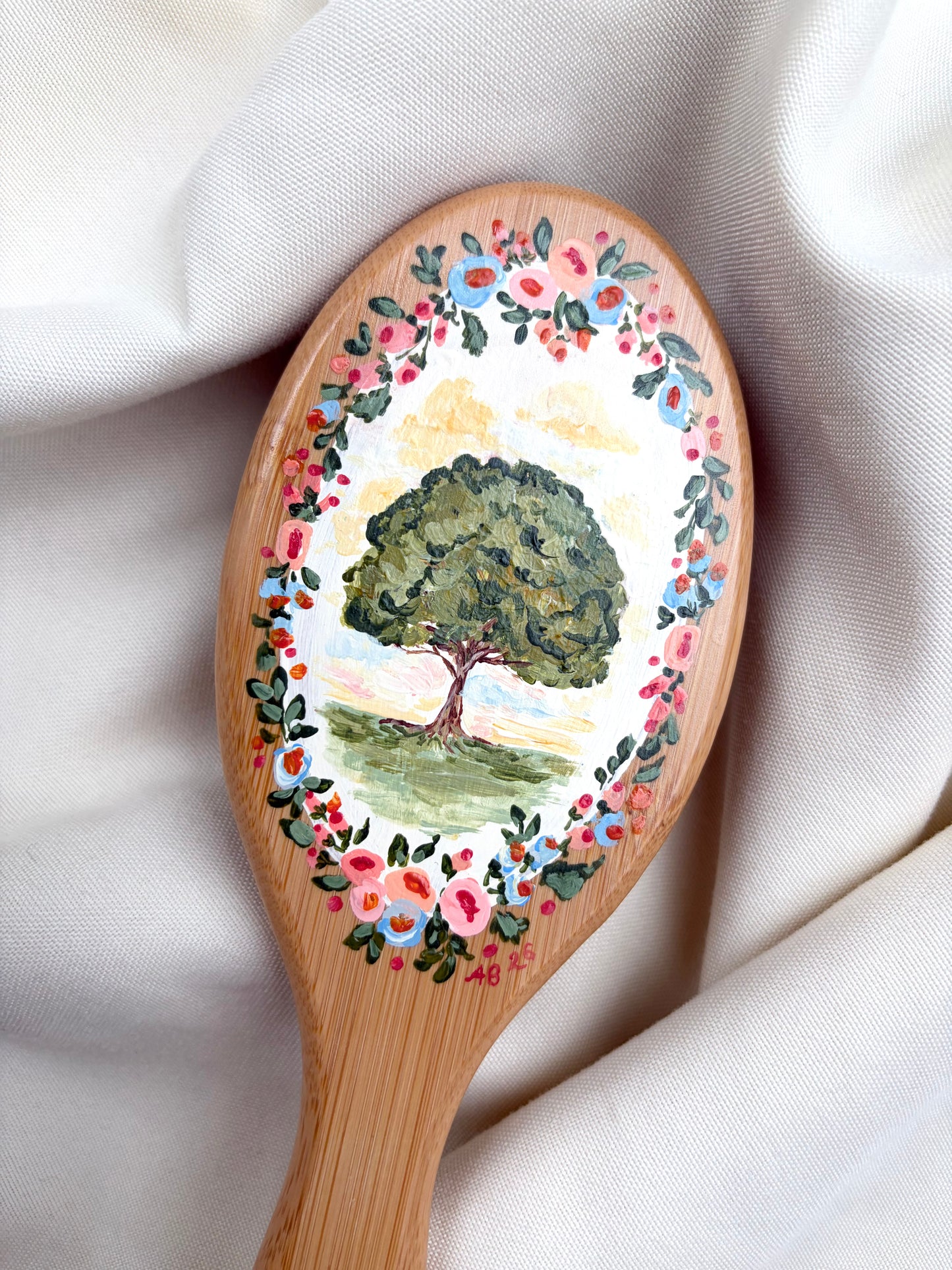 A close-up of a hairbrush handle with a hand-painted design featuring a green tree in the center and a border of pink, red, and blue flowers with green leaves, set against draped white fabric.
