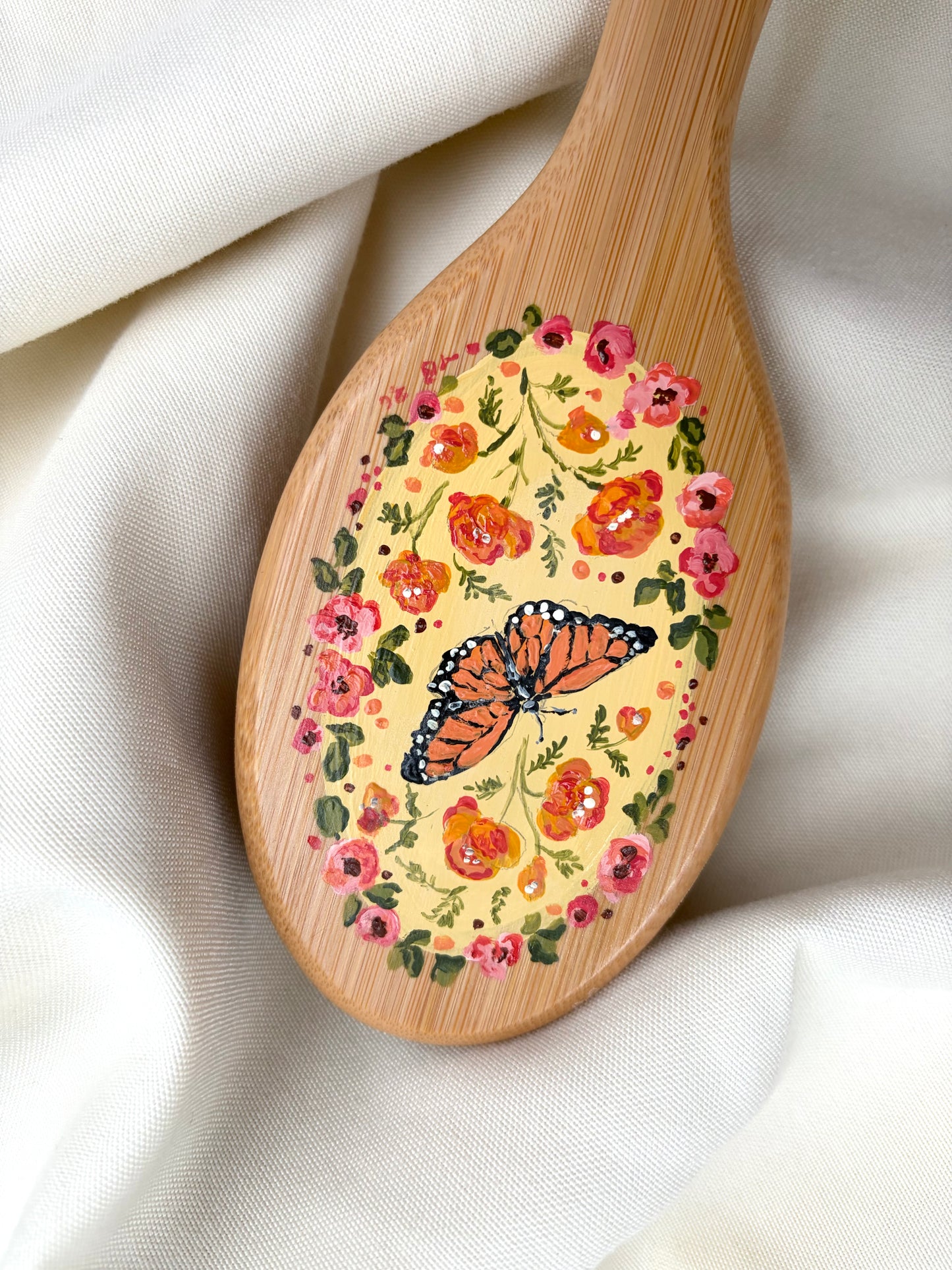 A close-up of a hairbrush painted with a colorful hand-painted design featuring a butterfly and a circle of flowers, resting on draped white fabric.