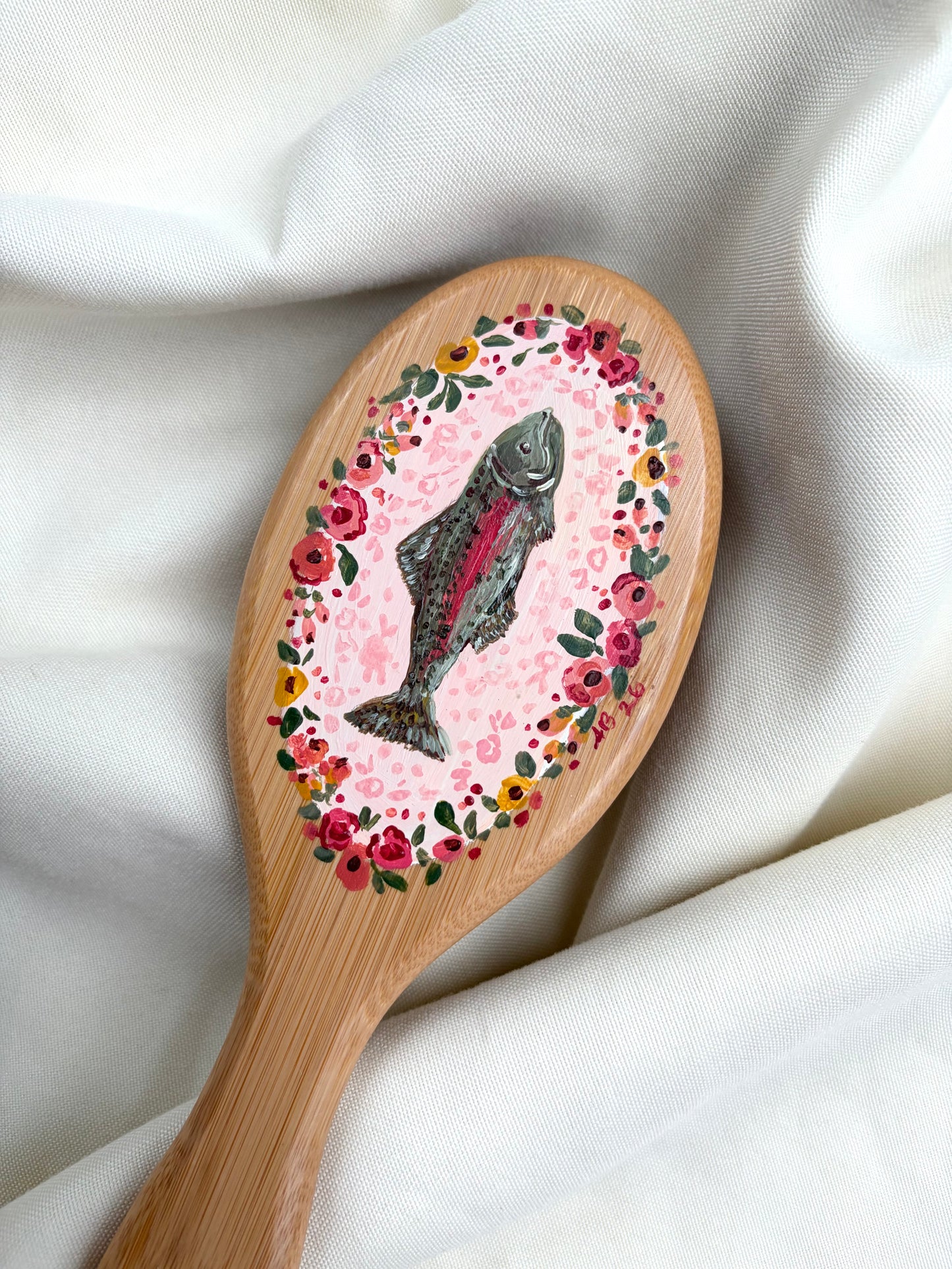 A wooden hairbrush with a hand-painted design featuring a fish in the center, surrounded by a pink floral border, resting on draped white fabric.