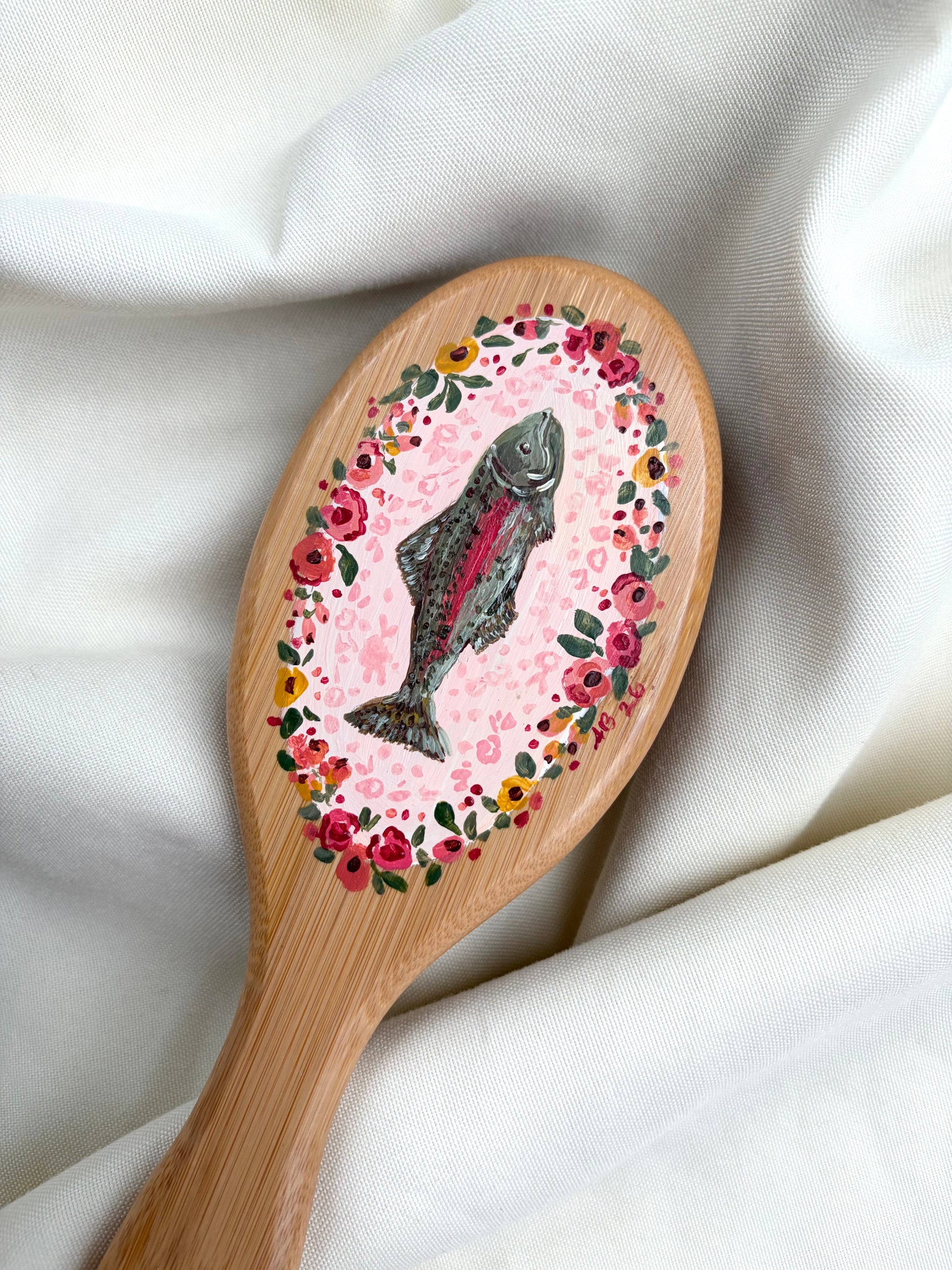 A wooden hairbrush with a hand-painted design featuring a fish in the center, surrounded by a pink floral border, resting on draped white fabric.